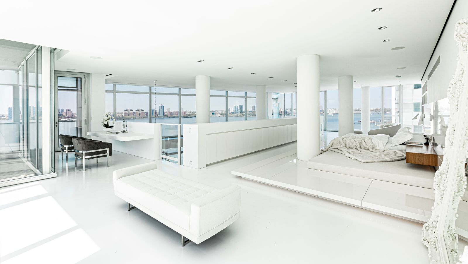 The sleeping area is set on a raised platform, slightly elevated from the surrounding floor, with the bed facing toward the windows and the city skyline. 