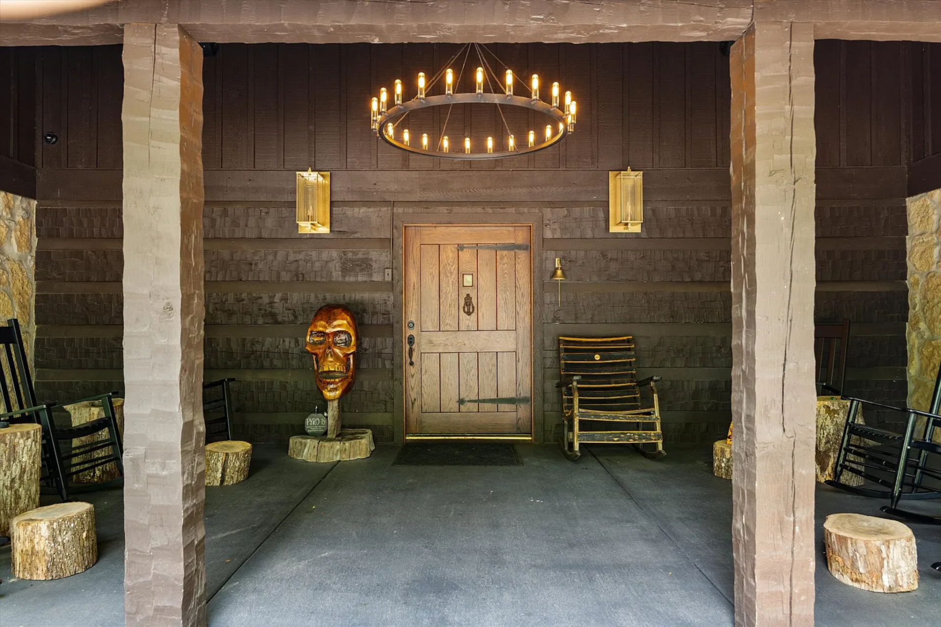 A deep covered porch frames the entrance with thick timber posts, dark wood siding, and rustic detailing, while the front door sits centered beneath a large circular chandelier.