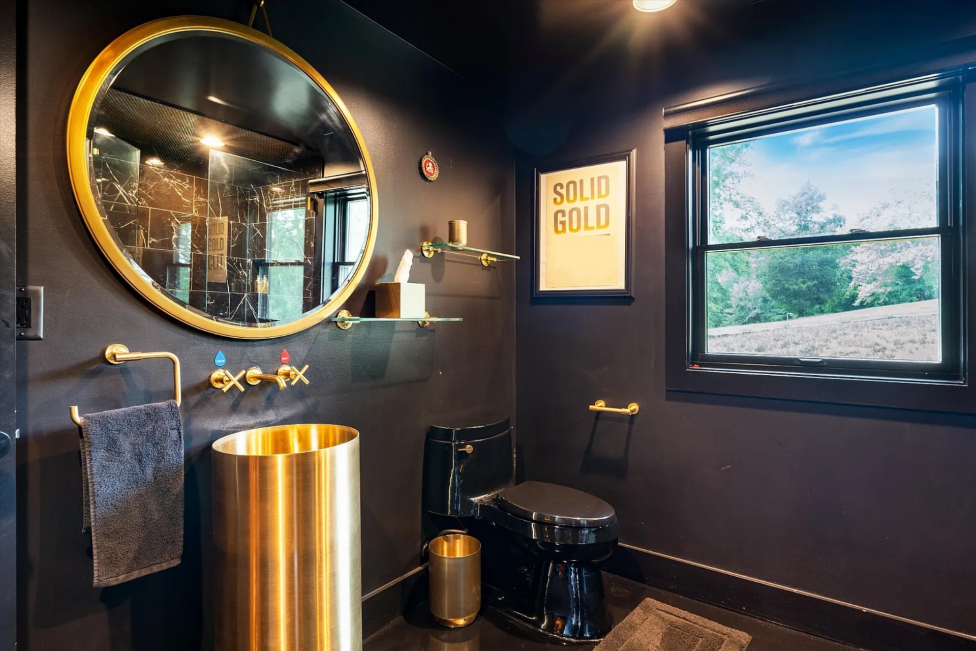 A cylindrical gold-toned sink and matching fixtures stand out against matte black walls, while a round mirror and compact layout keep the space efficient without feeling overly utilitarian.