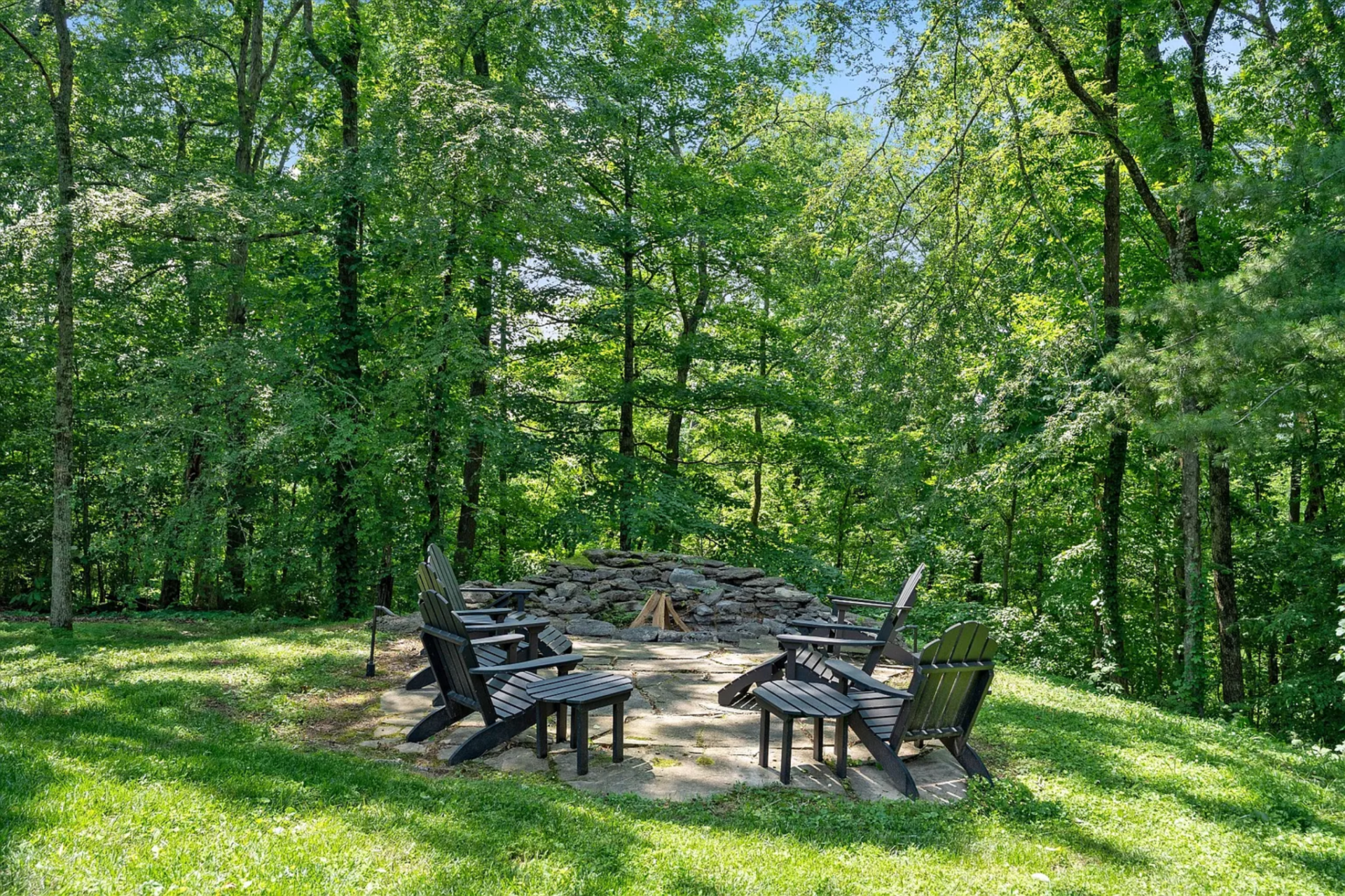 Positioned at the edge of the woods, this spot creates a natural gathering point, with a low stone wall framing the fire feature and the surrounding tree canopy enclosing the space.