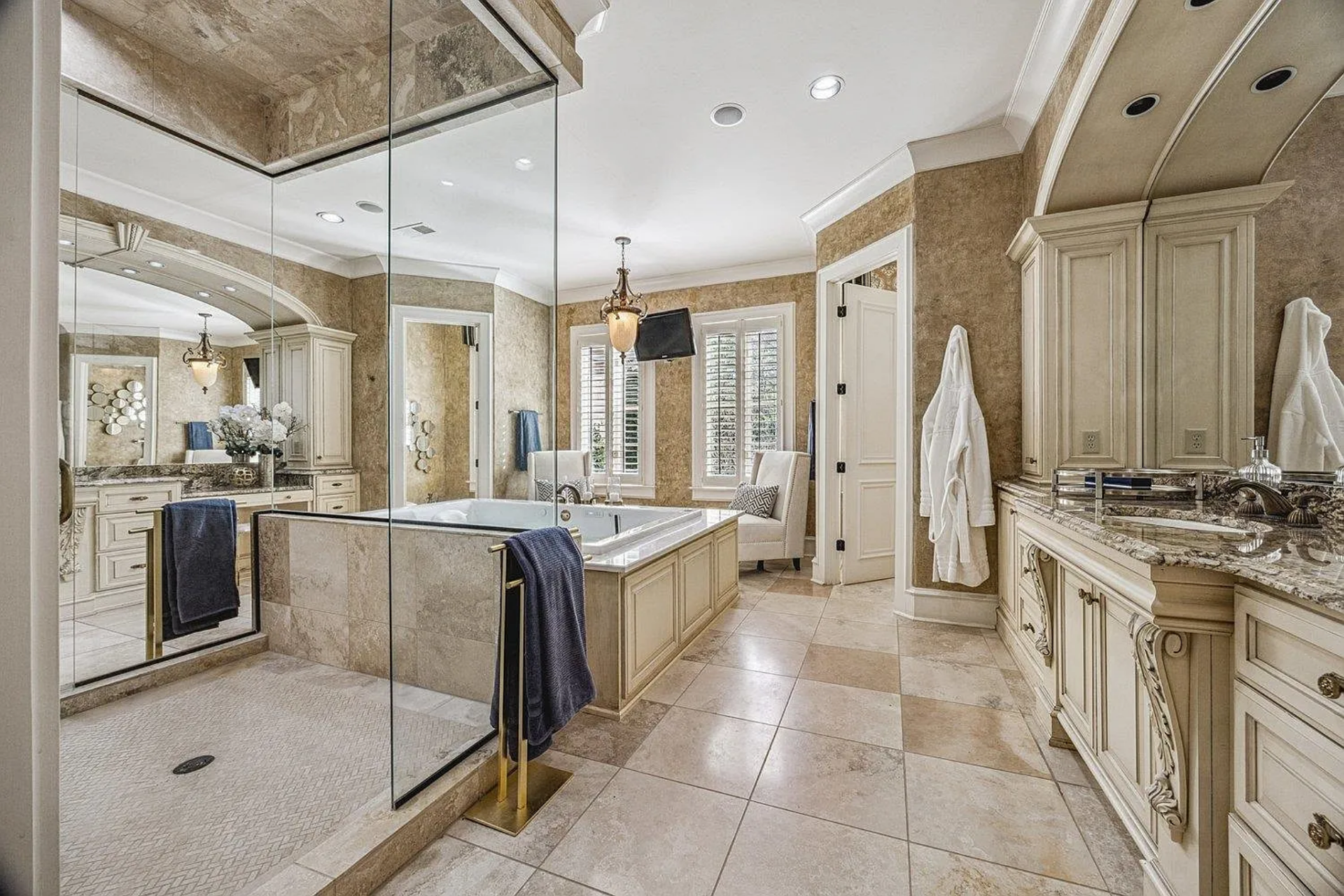 A glass-enclosed walk-in shower sits in the middle of the room behind the tub, while long vanities with carved cabinetry and stone countertops run along either side.