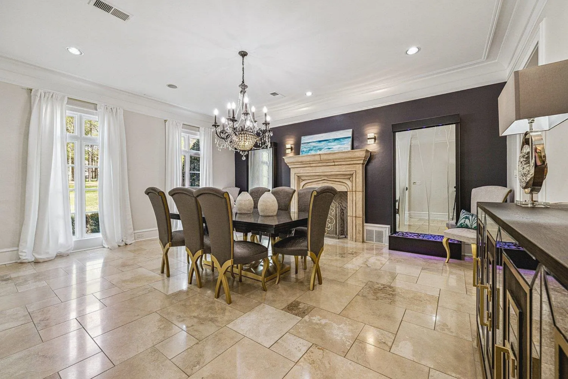 Around the corner, the formal dining room features a rectangular table surrounded by high-back upholstered chairs with gold-finished frames, positioned to face the fireplace along the far wall.