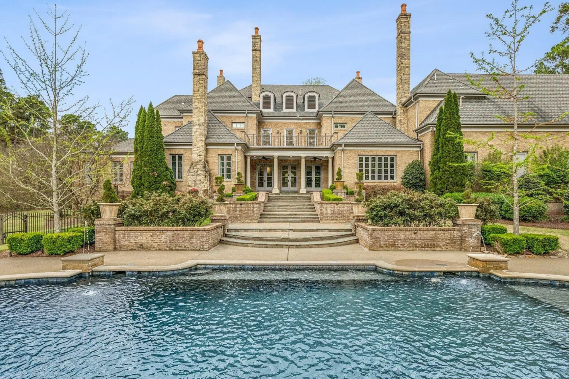 The pool itself serves as the focal point, framed by stone decking and accented with water features, while the rear of the home rises symmetrically, complete with multiple chimneys, terraces, and manicured landscaping that reinforce the estate’s scale and presence.