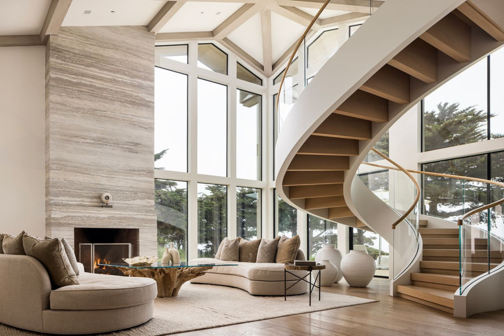 Beneath a curving staircase, the seating area is arranged with low, rounded sofas and a natural wood coffee table, keeping the room open while still defining it as a central gathering space.