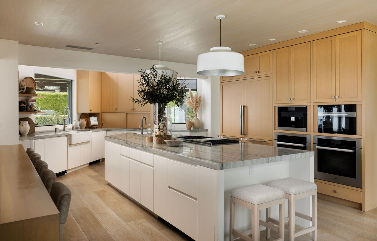 Near the dining area, the kitchen is organized around a large central island finished with a thick stone countertop, with two stools tucked at one end for casual seating. 