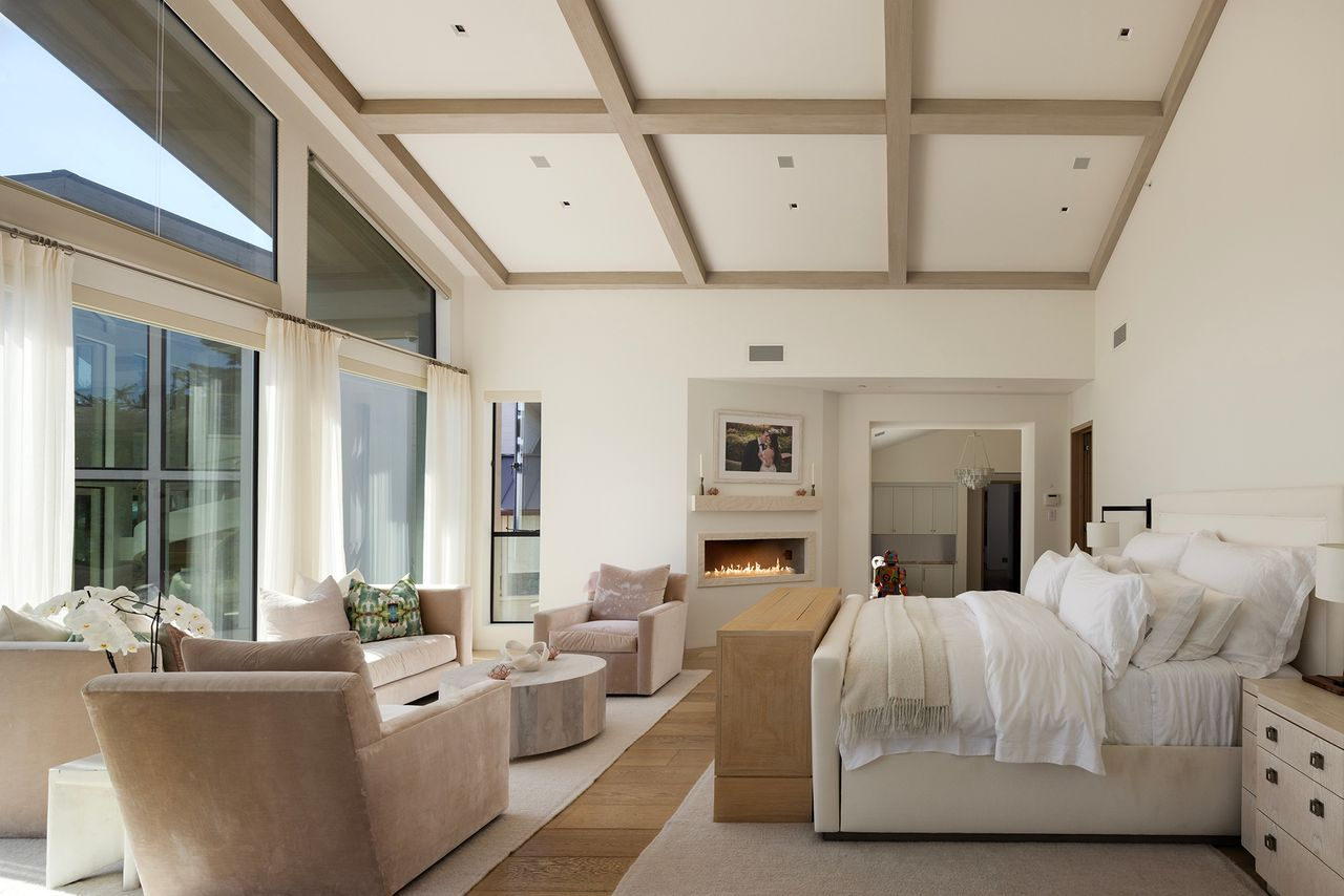The bed is positioned to face both the fireplace wall and the sitting area, with a pair of chairs and a low table set in front of the windows.