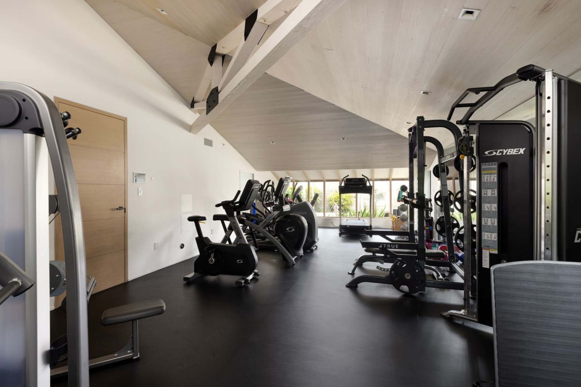 The gym is laid out as a full training room, with black flooring running wall to wall beneath a mix of cardio and strength equipment. 