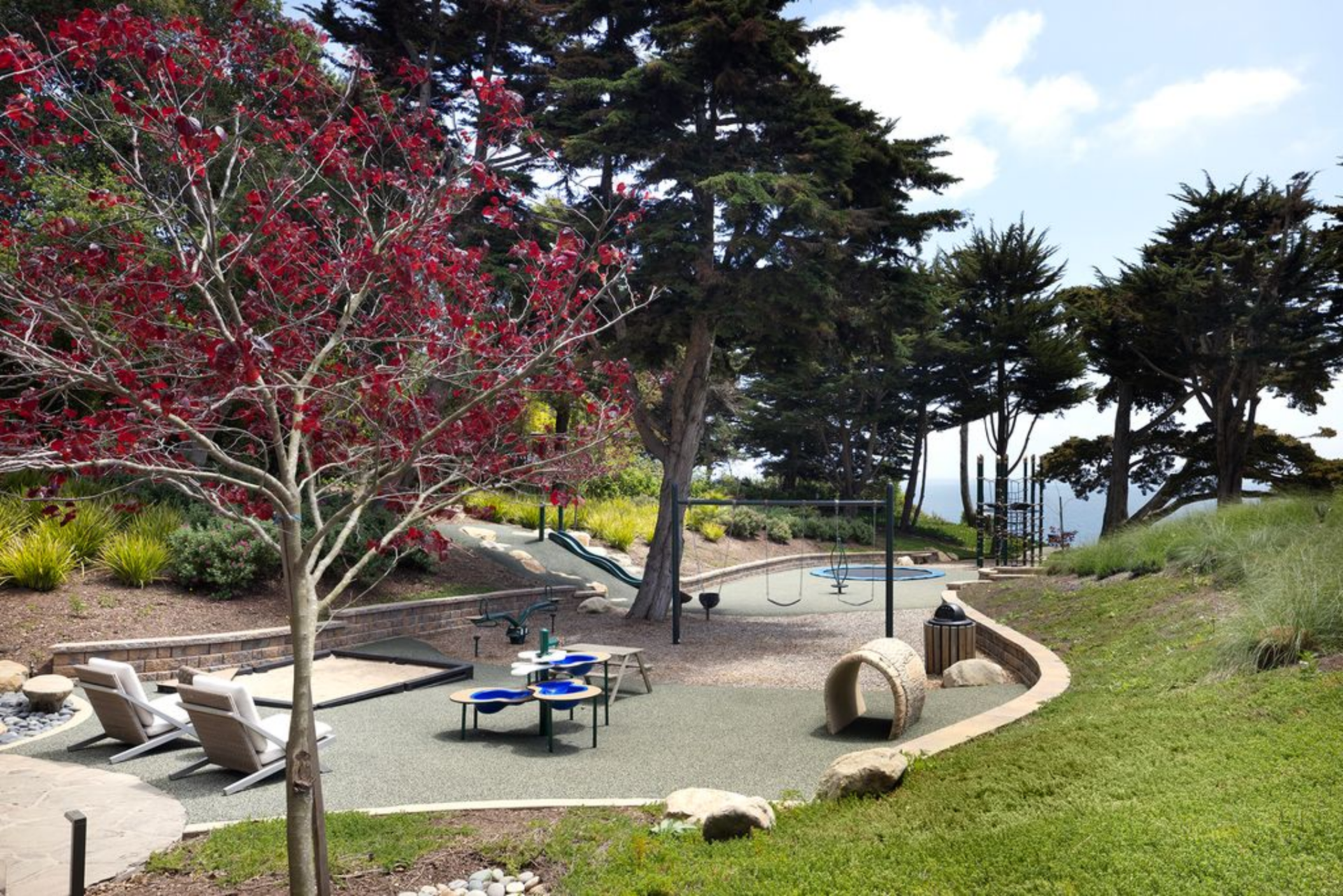 A mix of swings, climbing structures, a sandbox, and an in-ground trampoline are built into a recessed zone bordered by low retaining walls and tall trees.