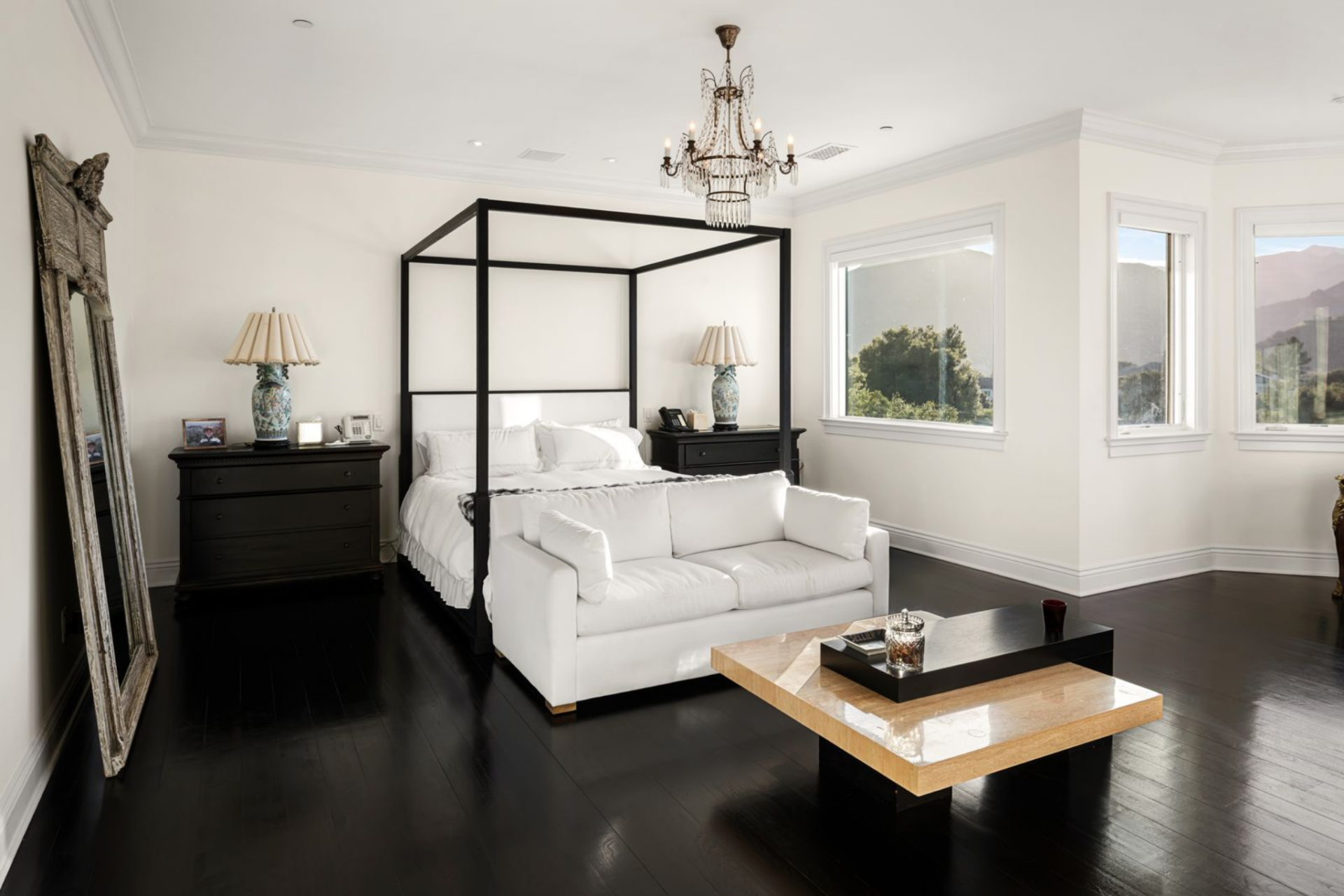 The bedroom centers around a four-poster bed positioned beneath a chandelier, with a sitting area placed at the foot to extend the room beyond just sleeping. 
