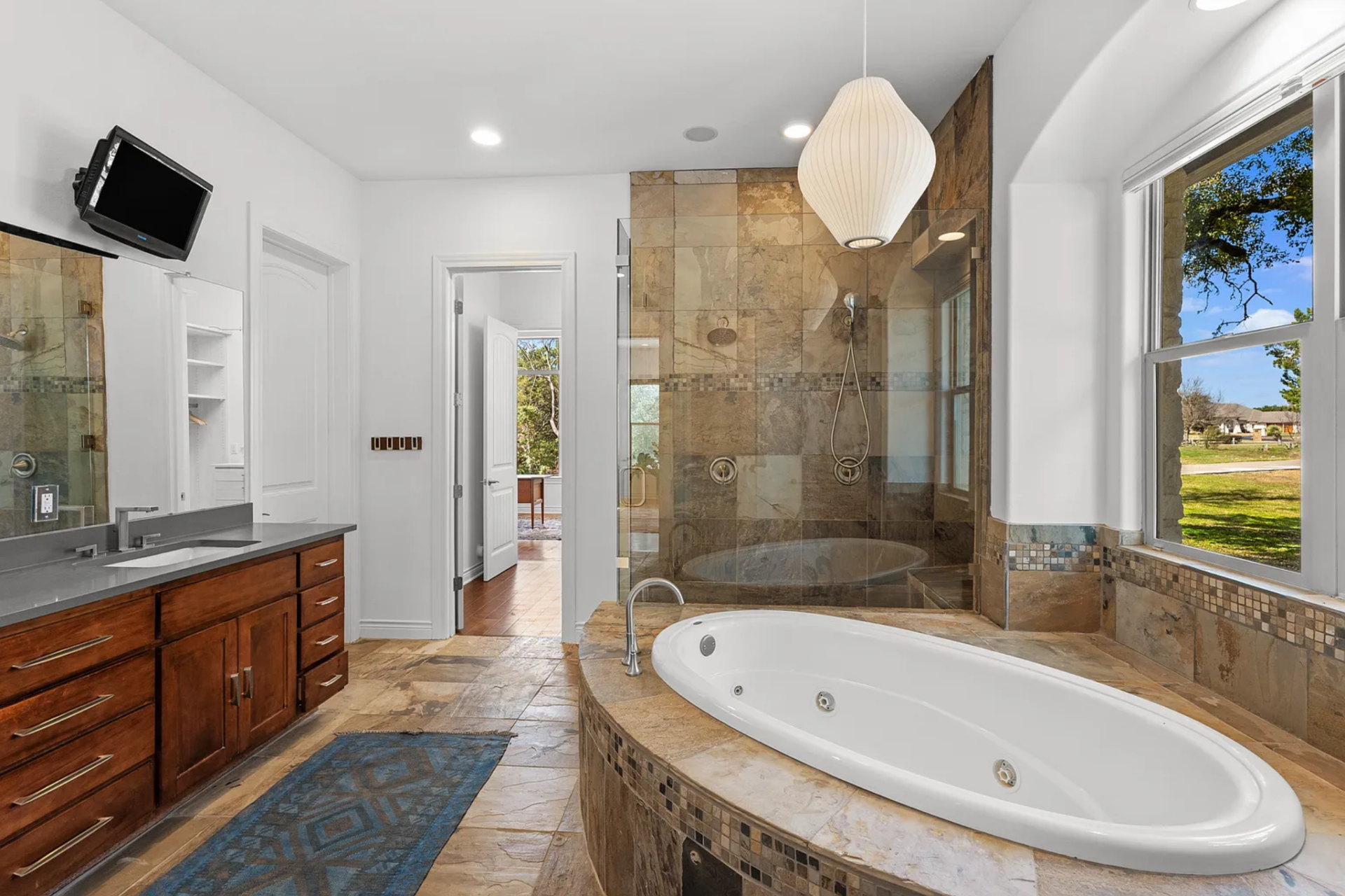 Opposite, a built-in soaking tub is set into a raised stone surround, positioned beneath a window and next to the glass walk-in shower.