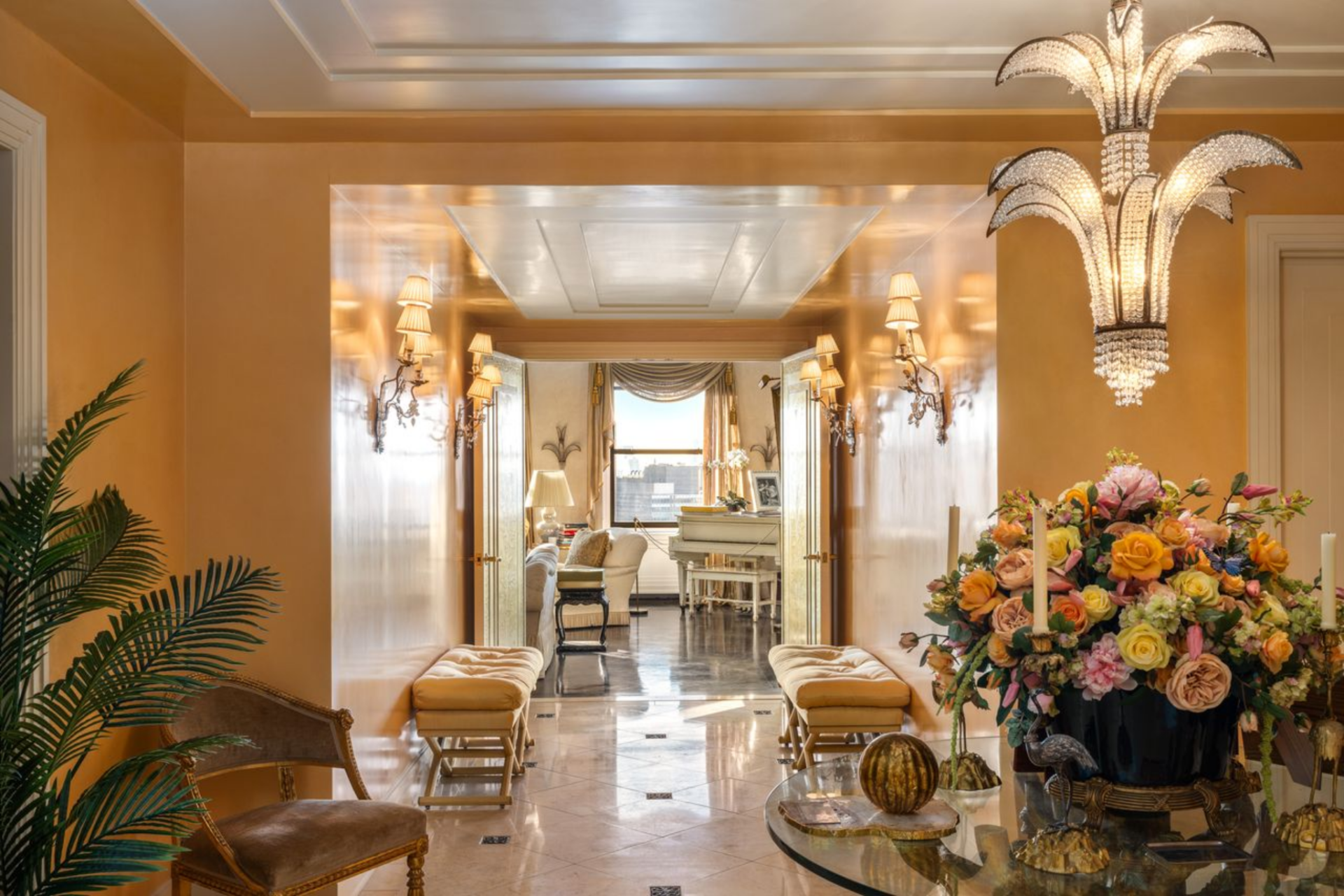 The space is wrapped in glossy peach-toned walls and centered on a crystal chandelier that drops down like a sculpture, while polished stone floors with dark inlaid accents give the long passage a formal, reflective finish.