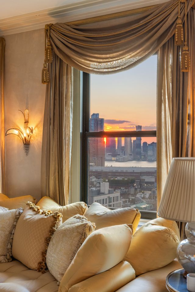 Along the perimeter, the windows frame wide city and river views, and in the closer view, one of those windows becomes a focal point on its own, dressed with heavy swag curtains and looking out toward the sunset over the skyline.