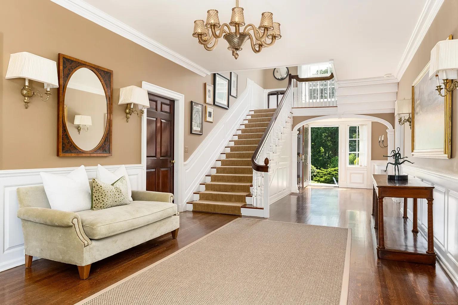 Stepping into the main house, a long entrance hall with dark wood floors run beneath a large neutral rug, while warm beige walls, white wainscoting, and layered crown molding give the space a traditional framework consistent with the home’s turn-of-the-century origins.