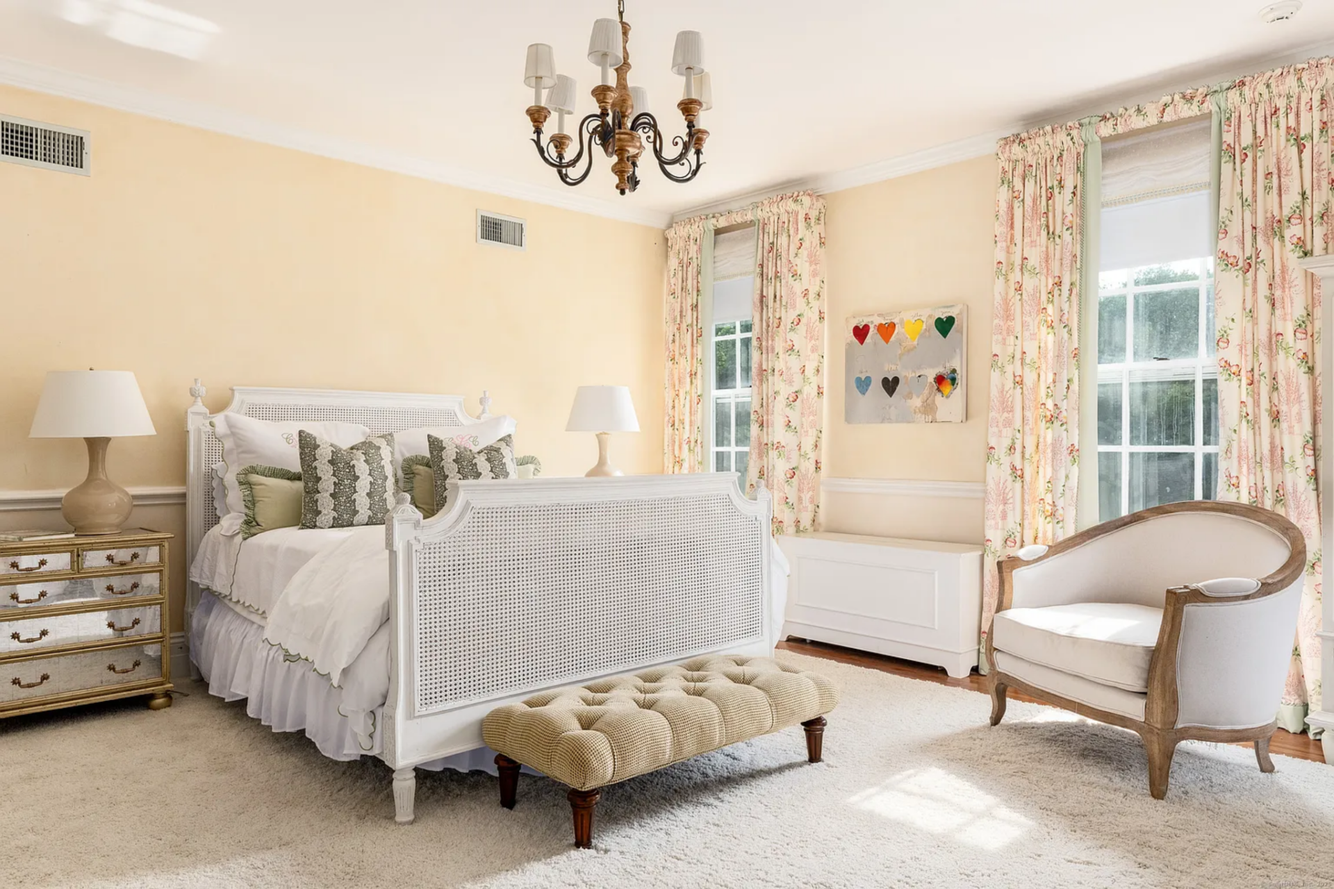 The next room has a warmer palette, with pale yellow walls, floral drapery, and a white cane-style bed set against the back wall. 