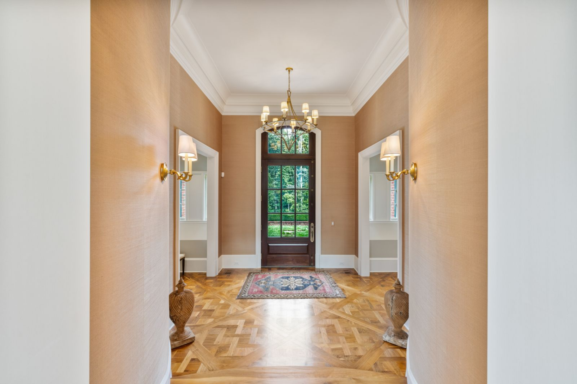 Walking into the main house, tall ceilings rise above a formal foyer wrapped in warm textured wallcoverings, while white trim and crown molding sharpen the architecture and frame the room with clean, traditional lines.