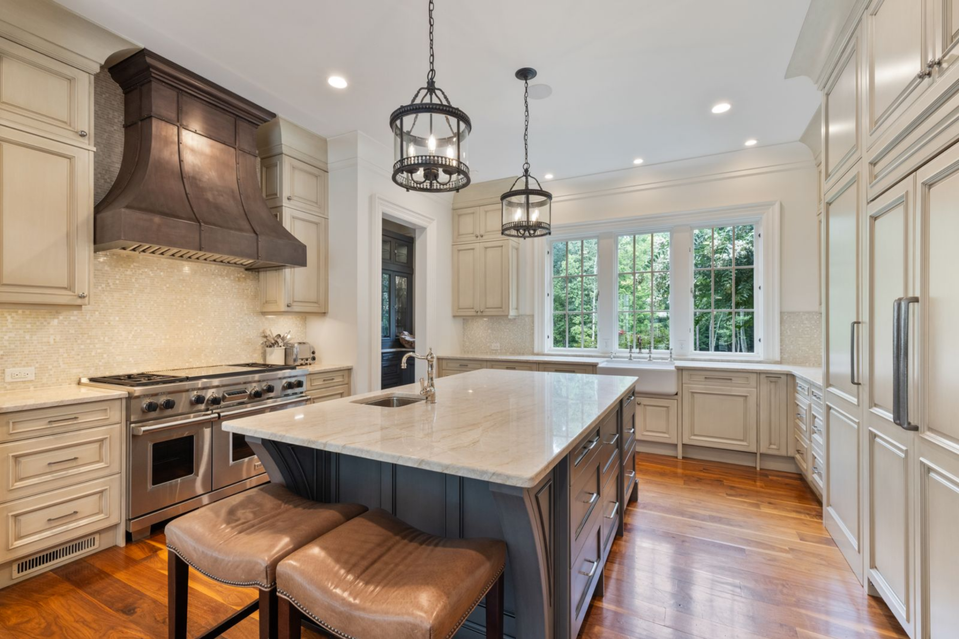 The island is finished in a darker tone than the surrounding cabinetry, which helps define it as the main prep and gathering point, with two leather stools tucked along one side for quick seating.