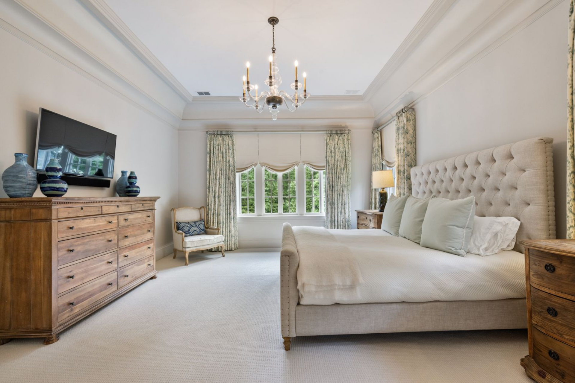 The bedroom itself is arranged with the bed centered along one wall beneath a tall upholstered headboard, while three windows bring in natural light and frame the greenery outside. 