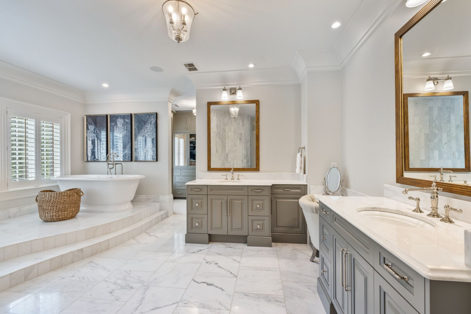 Off to one side, a freestanding soaking tub sits raised on its own platform beneath shuttered windows, while two separate vanities run along the other with painted cabinetry, framed mirrors, and white counters. 