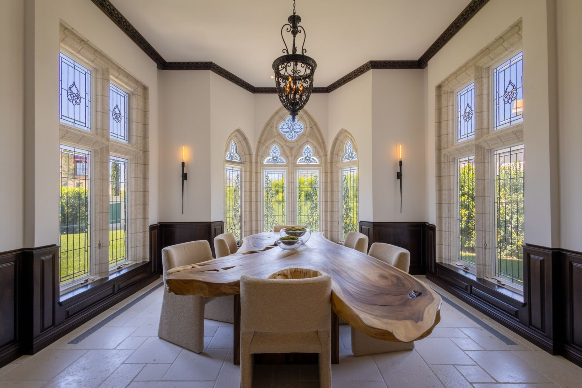 At the center, a live-edge wood table introduces a warmer, more organic element against the carved stone and dark paneled wainscoting, surrounded by upholstered dining chairs that soften the room.