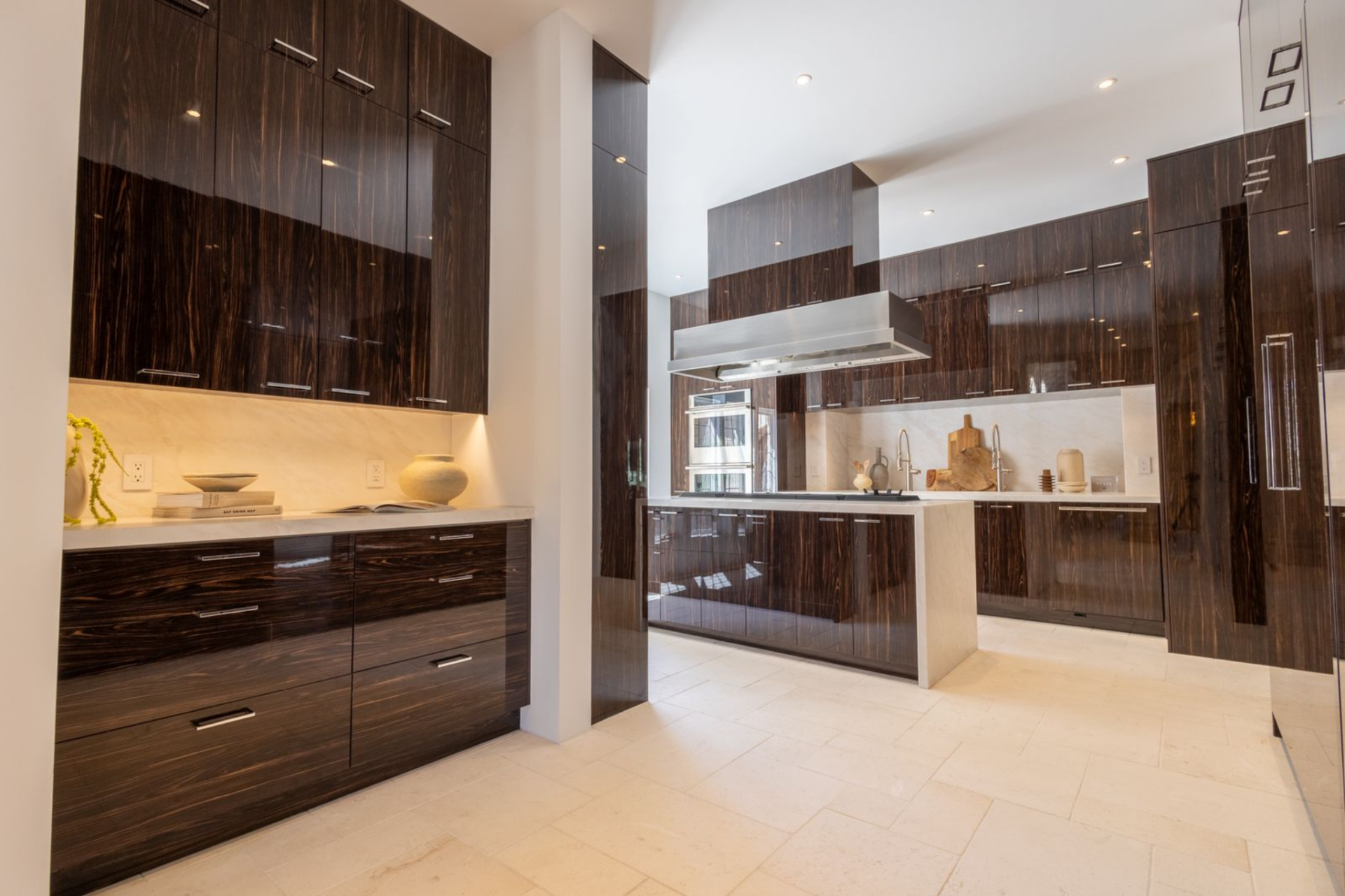 Tall glossy cabinetry in a deep espresso finish wraps the walls almost from floor to ceiling, giving the space a polished look, while pale stone floors and matching countertops keep the overall palette bright.
