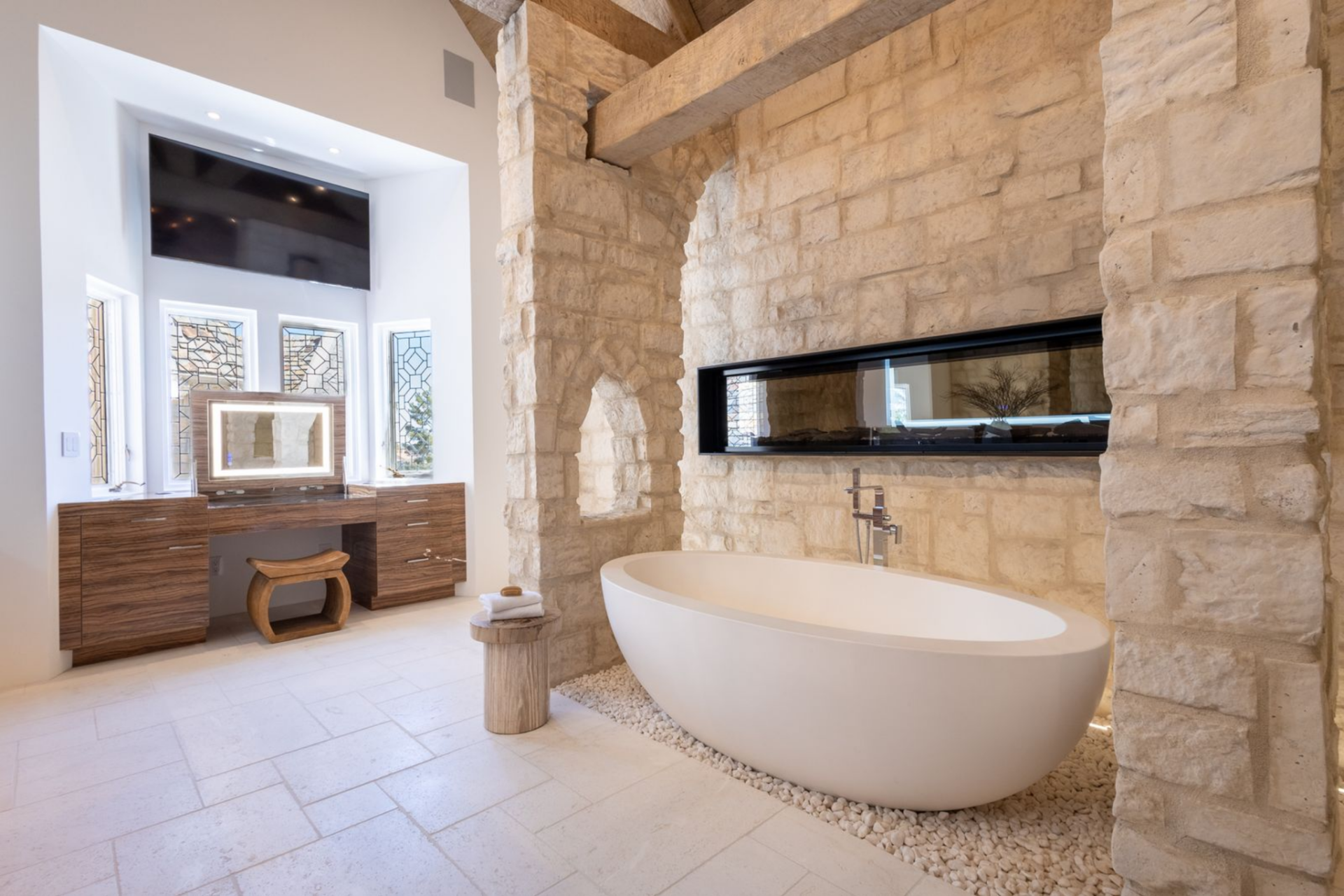 In the bathroom, a freestanding tub sits in front of a limestone wall and beneath exposed wood beams, with a long horizontal fireplace cut directly into the stone behind it.