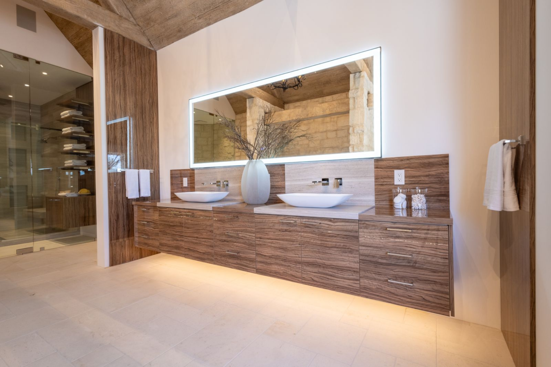 Across from the tub, a long floating vanity in richly grained wood stretches beneath a large backlit mirror, with vessel sinks set on top rather than recessed into the counter.
