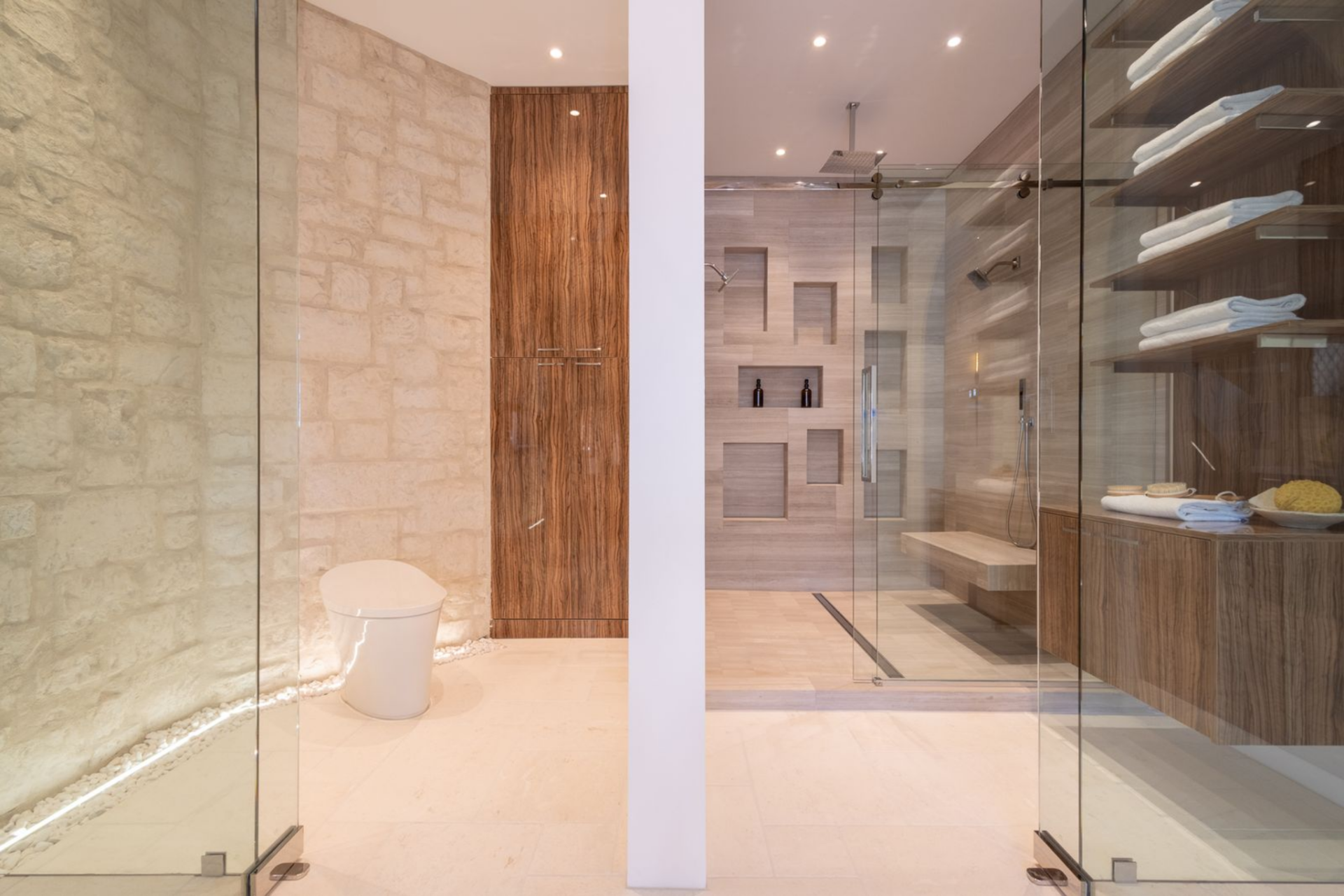 Off to the side, the glass-enclosed shower area features matching pale stone floors, built-in wood shelving, and a bench that reinforces the suite’s spa-like layout.