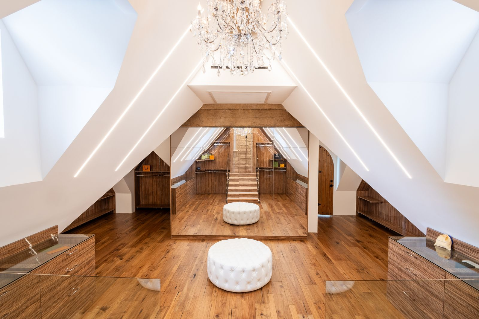 The suite's closet sits at the top of a short spiral staircase and under steep rooflines opening into a symmetrical dressing area with warm wood floors, built-in wood cabinetry, and mirrored central storage that reflects the full height of the space.