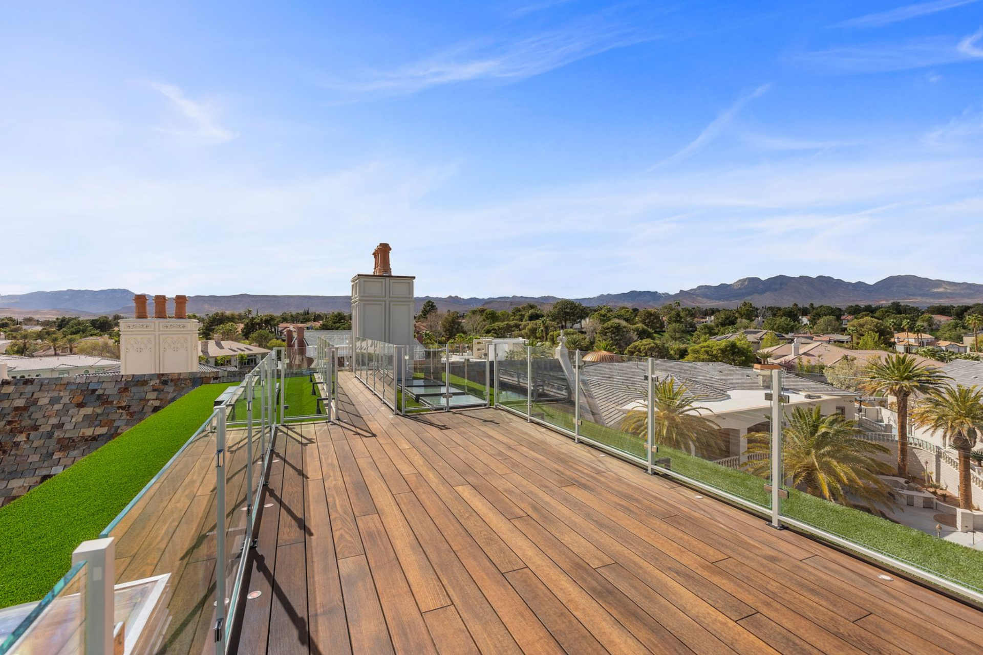 At the top, the rooftop deck functions as an open-air lookout above the house, finished with wide-plank decking, glass railings, and narrow strips of turf along the edges.