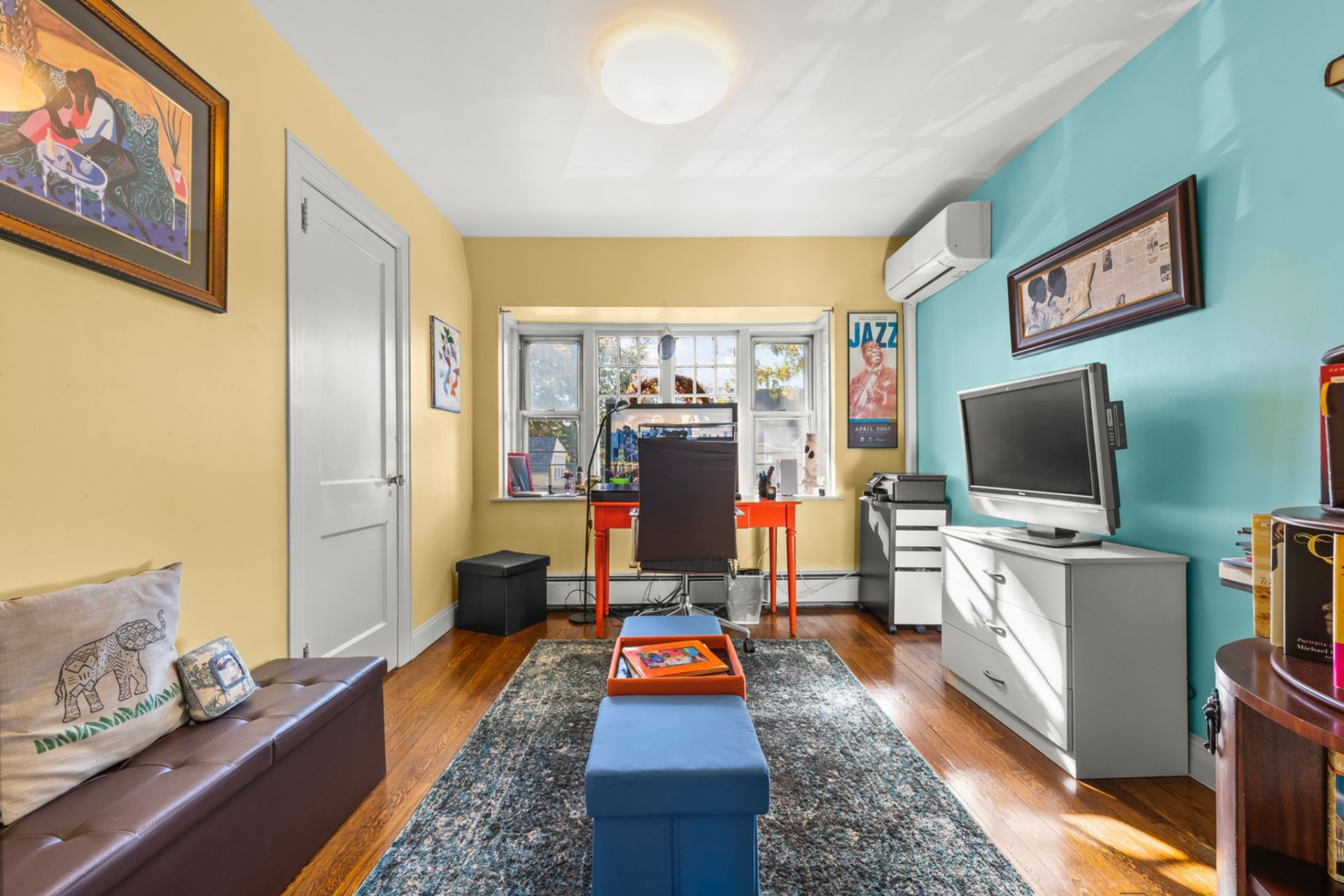 Inside, a desk is positioned in front of the window, with storage and a secondary seating area across from a TV, allowing the workspace to remain separate while still easily accessible from the bedroom.