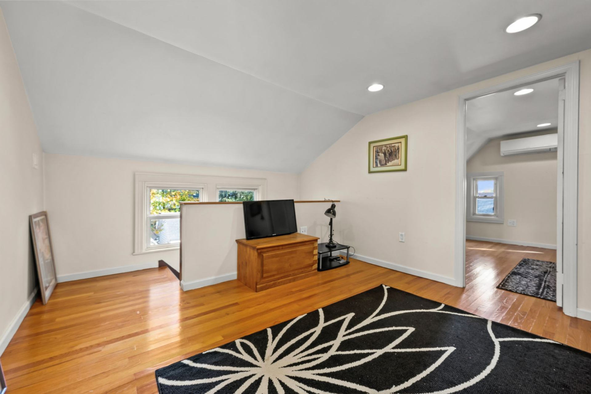 The room opens with a low half-wall overlooking the stairwell, and a small media setup is arranged along one side with a cabinet and TV. 