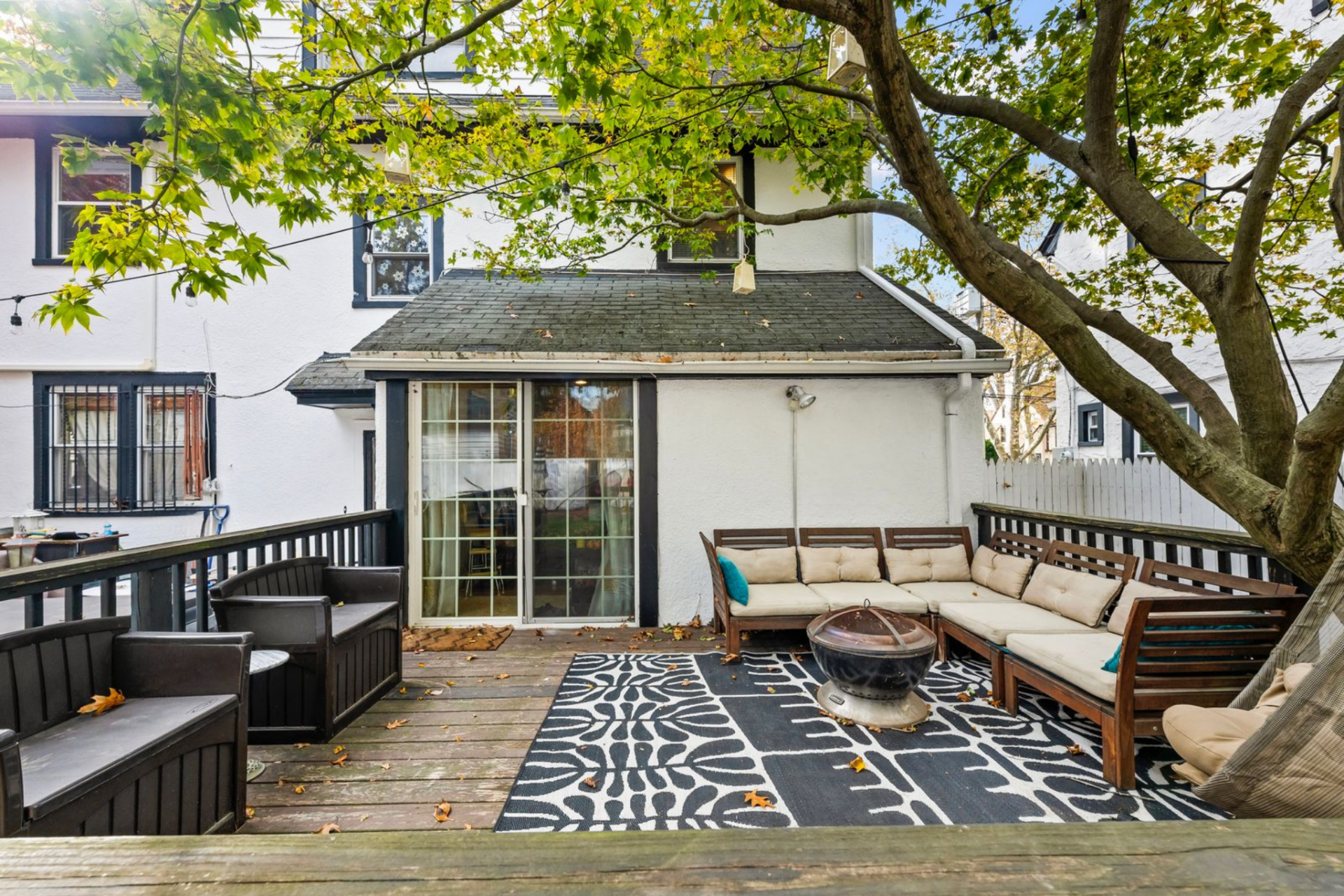 The space is arranged with bench seating, chairs, and a central fire pit, forming a defined gathering area with branches providing partial shade overhead.