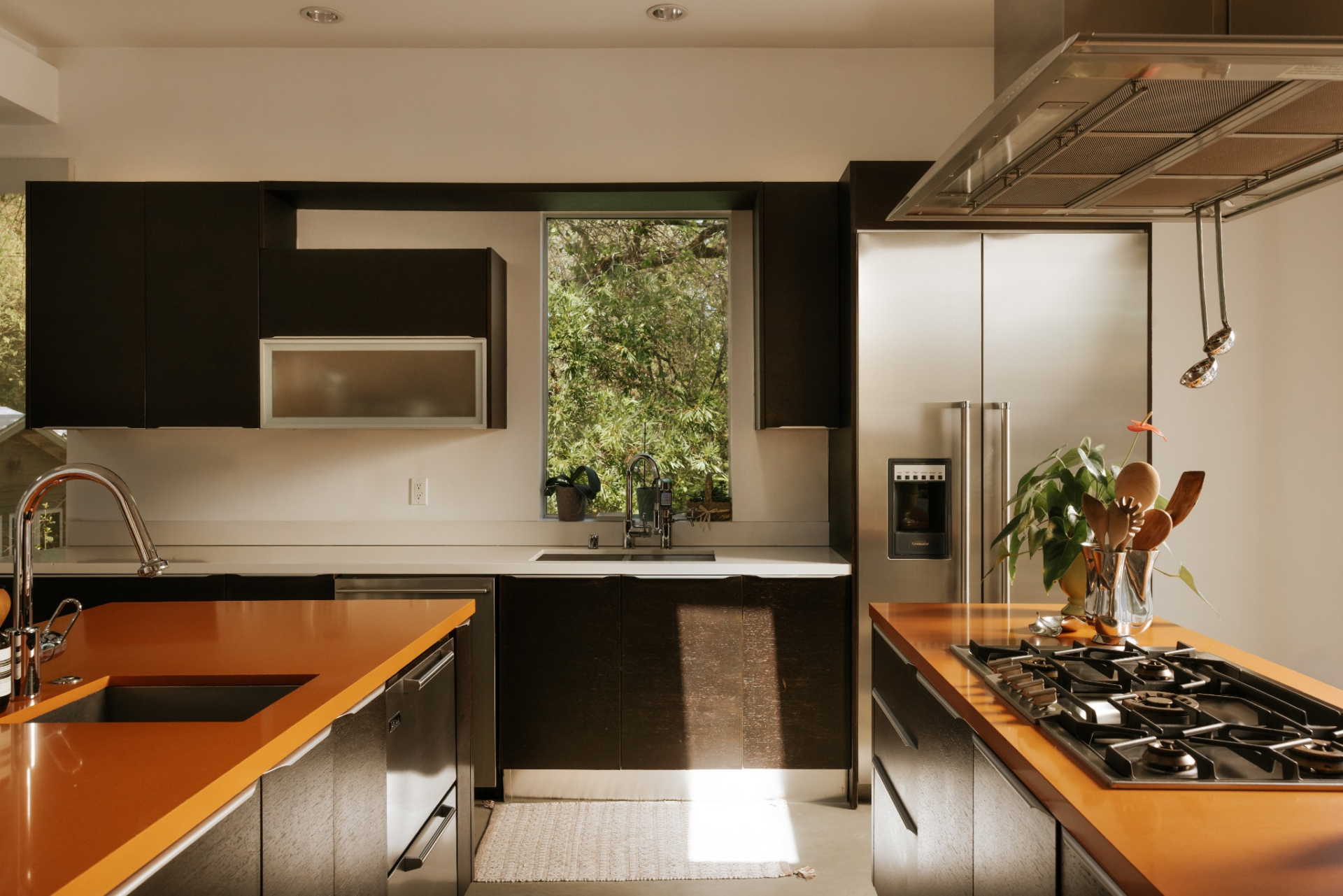 A window set above the main sink brings in natural light and frames greenery outside, while sliding glass doors just beyond the islands connect the kitchen directly to the outdoor area.