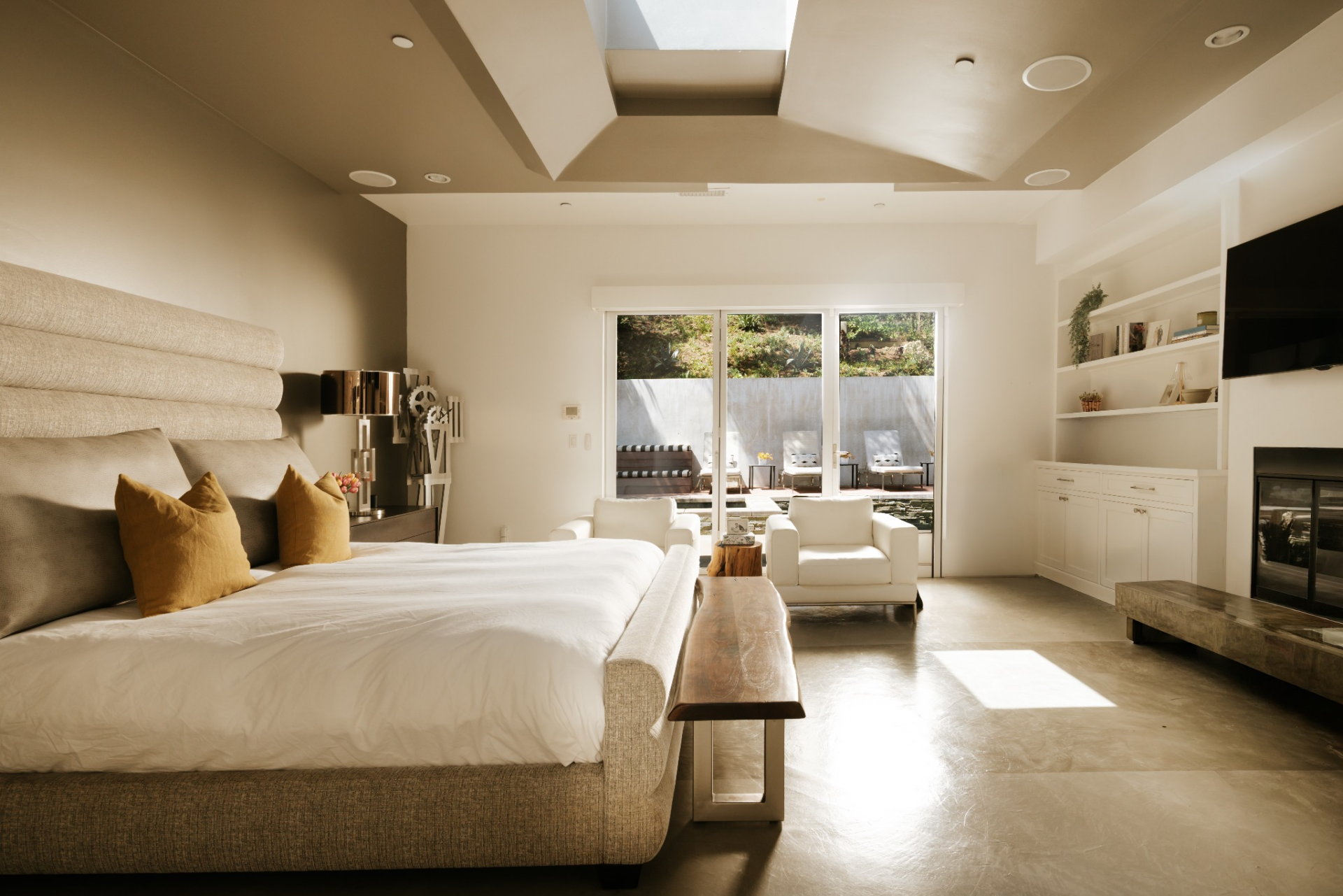 The bed sits at one end, a pair of chairs creates a small lounge in the middle, and the opposite wall incorporates built-in shelving, a fireplace, and a mounted television.