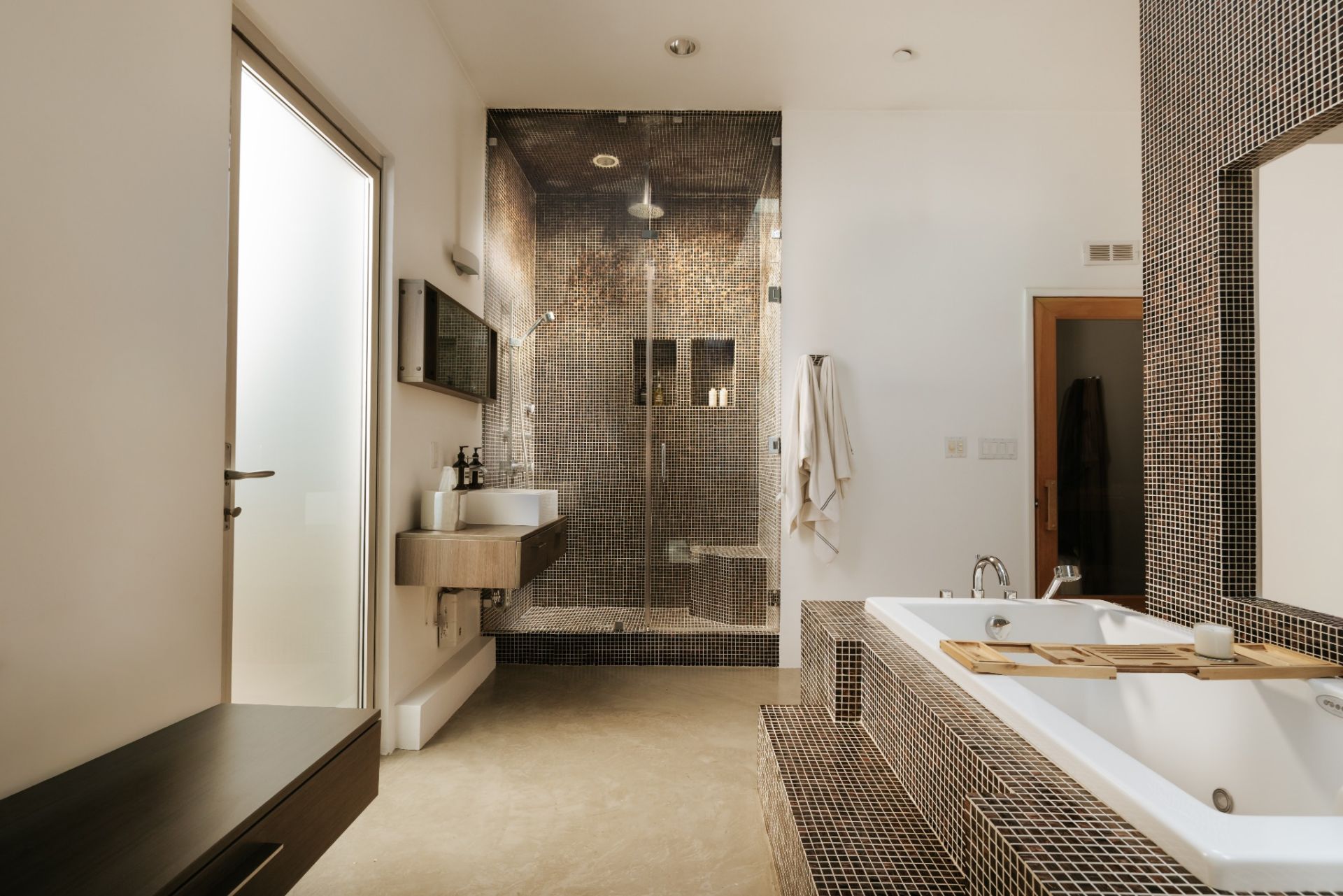 A deep soaking tub is built into a raised surround finished in small dark mosaic tile, while there is a glass-enclosed shower across the room.