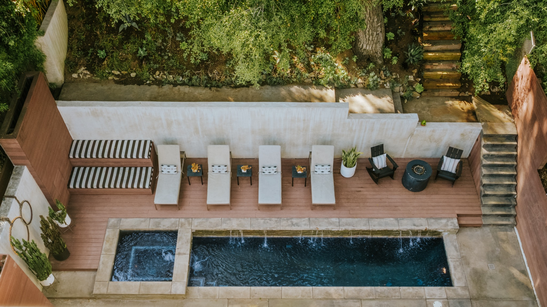 On one side, a raised wood deck lines the pool with a row of chaise lounges, built-in bench seating, and a small fire feature, while mature trees, planted slopes, and layered greenery rise around the perimeter.