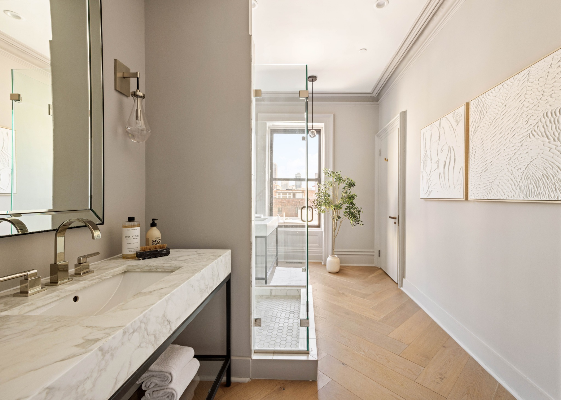 At the back of the bedroom, a door leads to the bathroom, which is arranged as a long, narrow space that places the glass-enclosed shower between two vanities.
