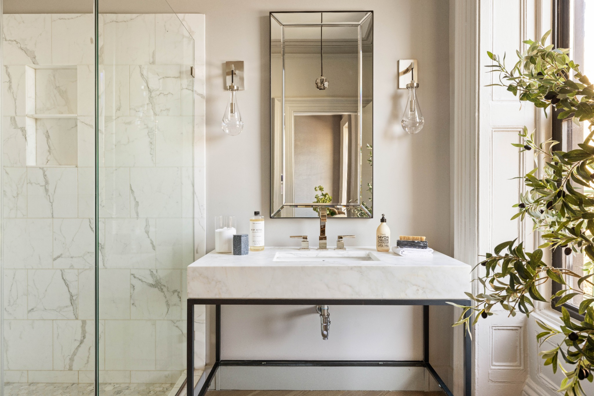 The vanities feature marble countertops and integrated sinks that sit on a slim metal base, while wall-mounted fixtures and mirrors maintain the clean, pared-back look.