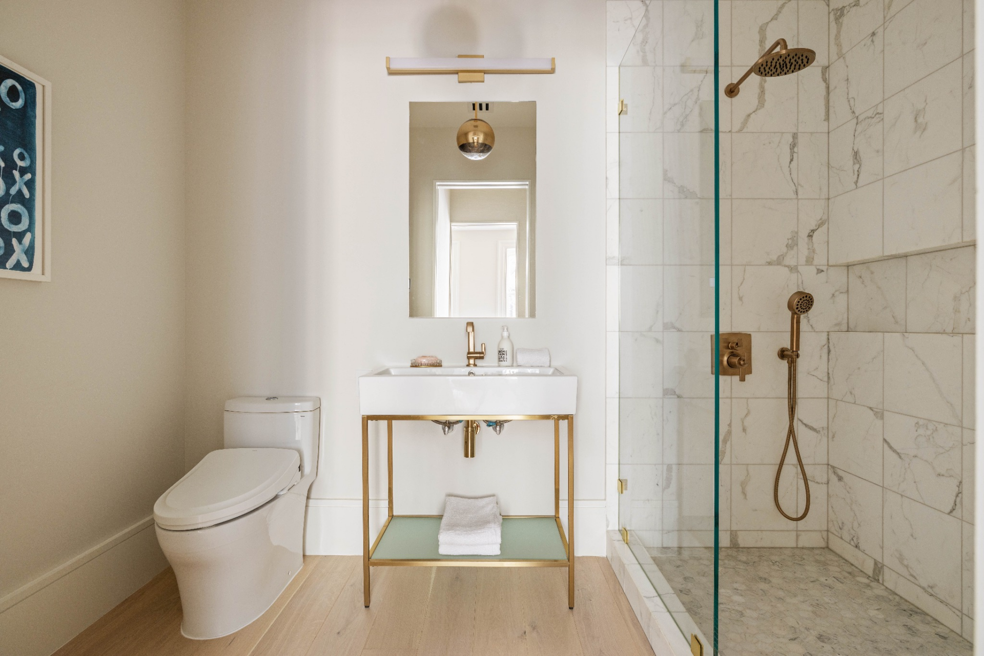 A slim vanity with exposed metal framing sits next to a glass-enclosed shower, both finished in marble and warm metallic fixtures.