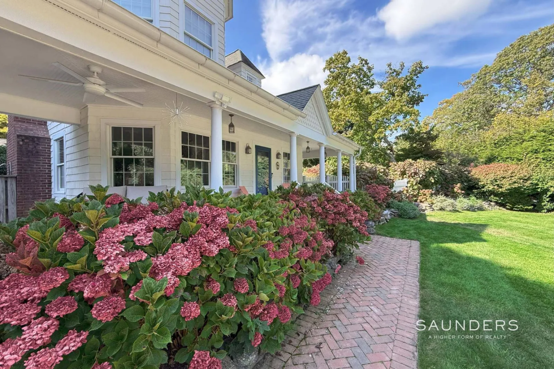 A brick path runs alongside thick hydrangea beds and opens to a deep stretch of lawn just before the round white columns that frame the porch. 