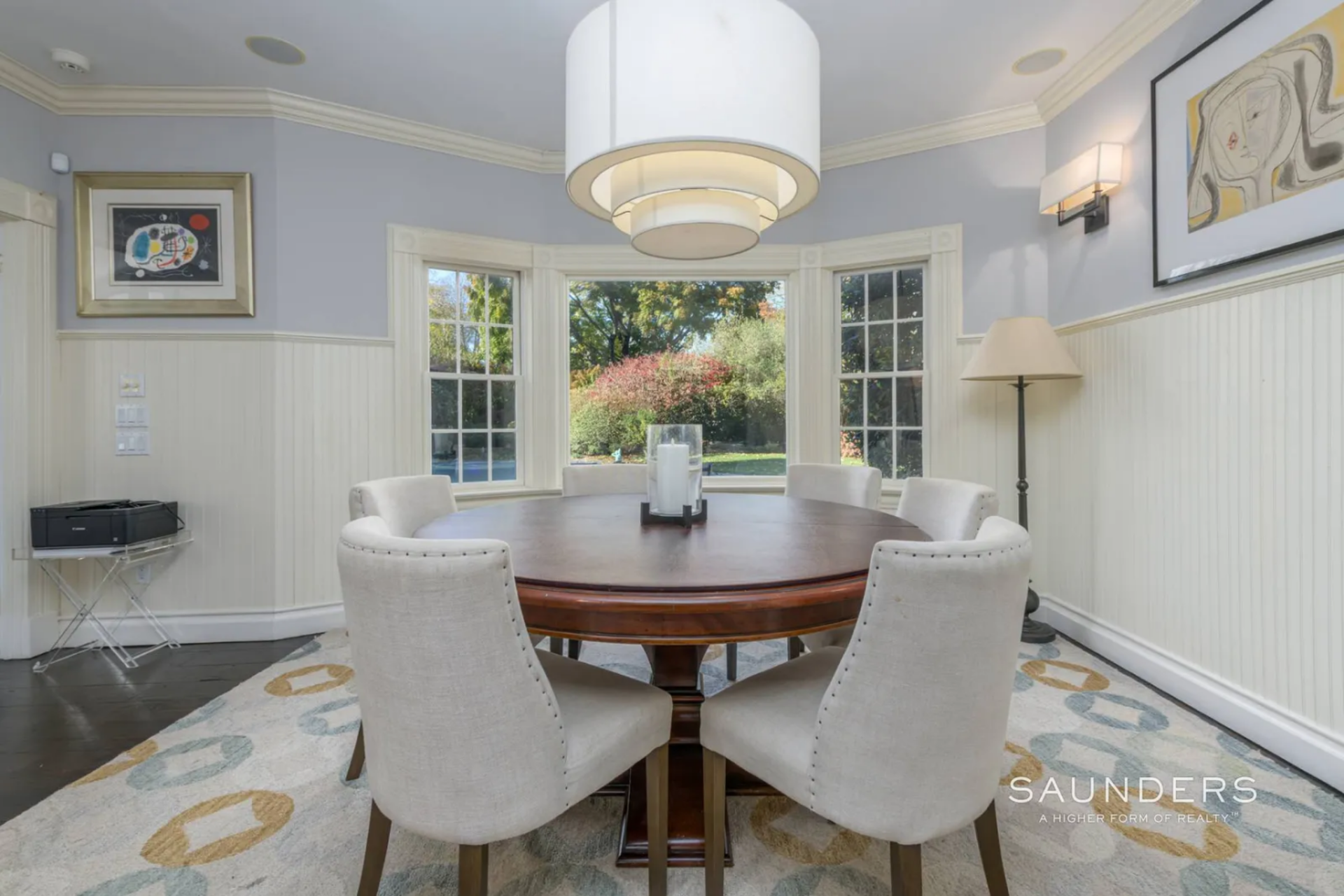 A round wood table sits at the center of the space beneath a large pendant light, with upholstered chairs placed evenly around it.