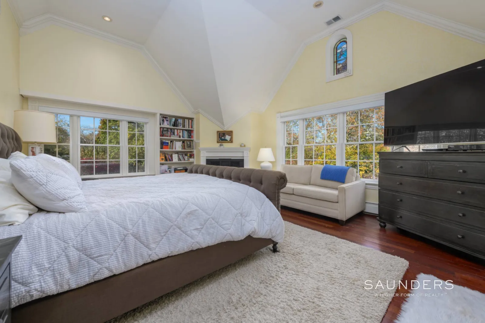 The bed sits at the center, framed by wide windows on two sides, while a fireplace is built into the far corner with shelving tucked beside it. 