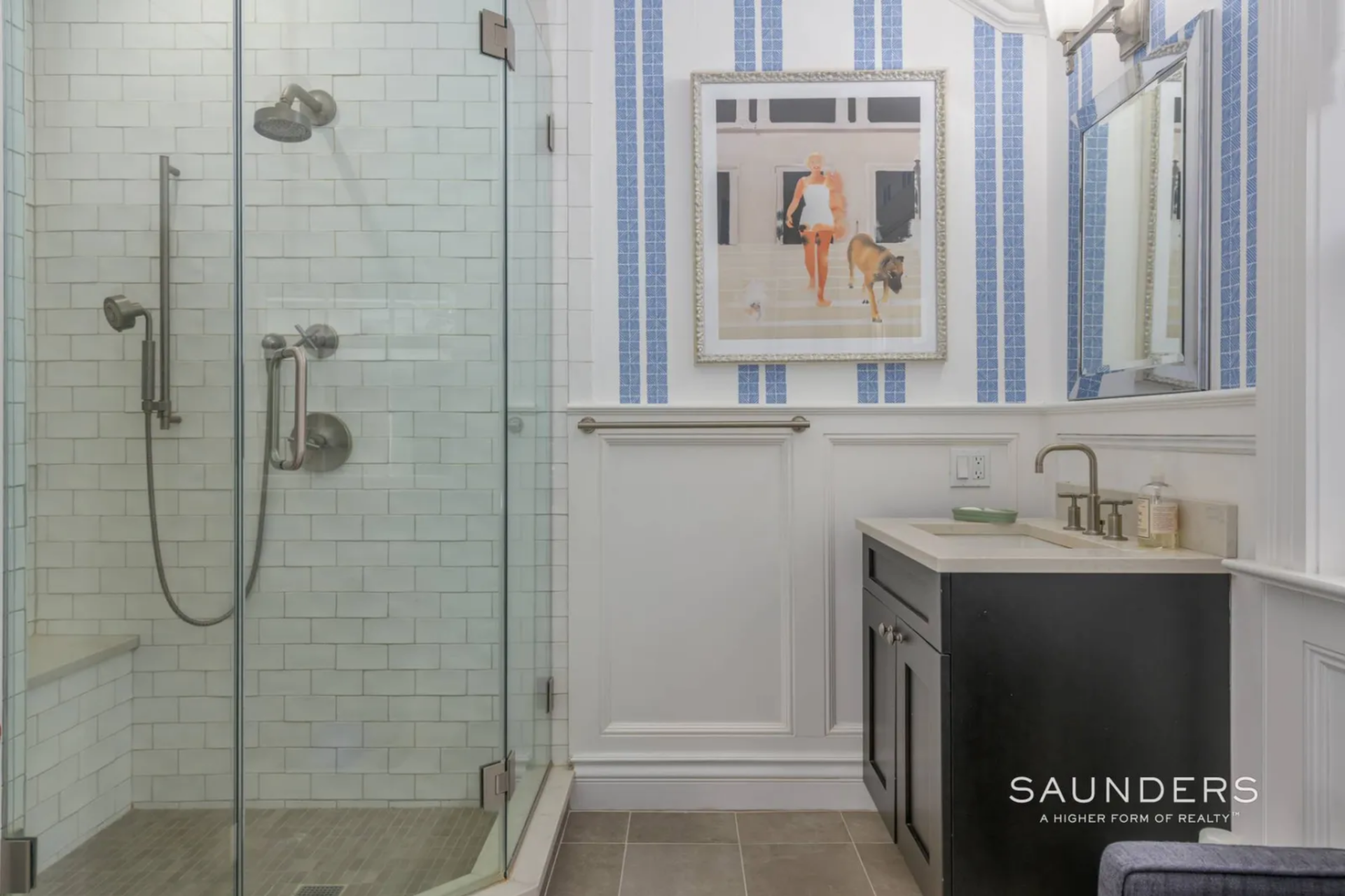 A glass-enclosed shower lines one side, finished in white subway tile, while the opposite side holds a dark vanity and paneled walls wrapped with blue vertical stripes that bring in a more decorative element.