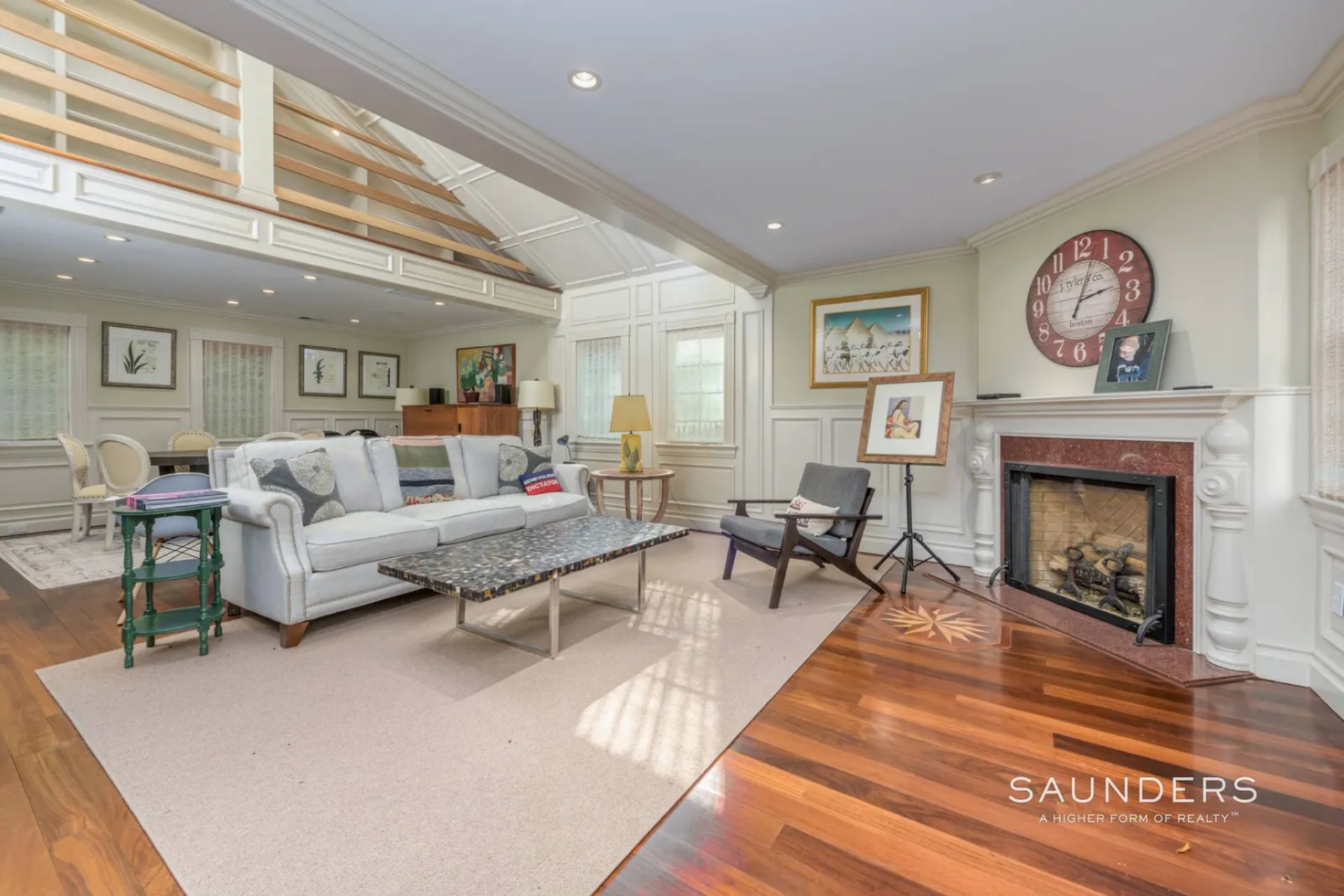 A fireplace sits on one side of the room, framed by a traditional mantel and surrounded by wall paneling, while the seating area is arranged openly in front of it with clear sightlines into a dining space at the far end.