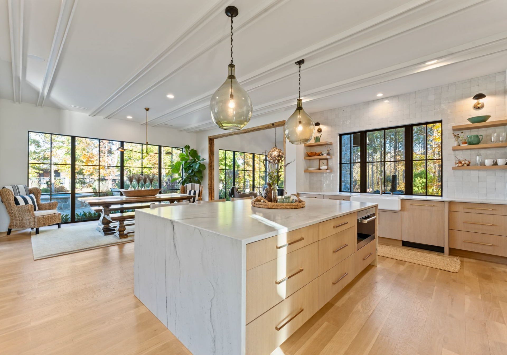 Quartzite surfaces run across the island and countertops, while light wood cabinetry keeps the palette soft, and the farmhouse sink, open shelving, and full wall of integrated storage balance the more minimal elements.