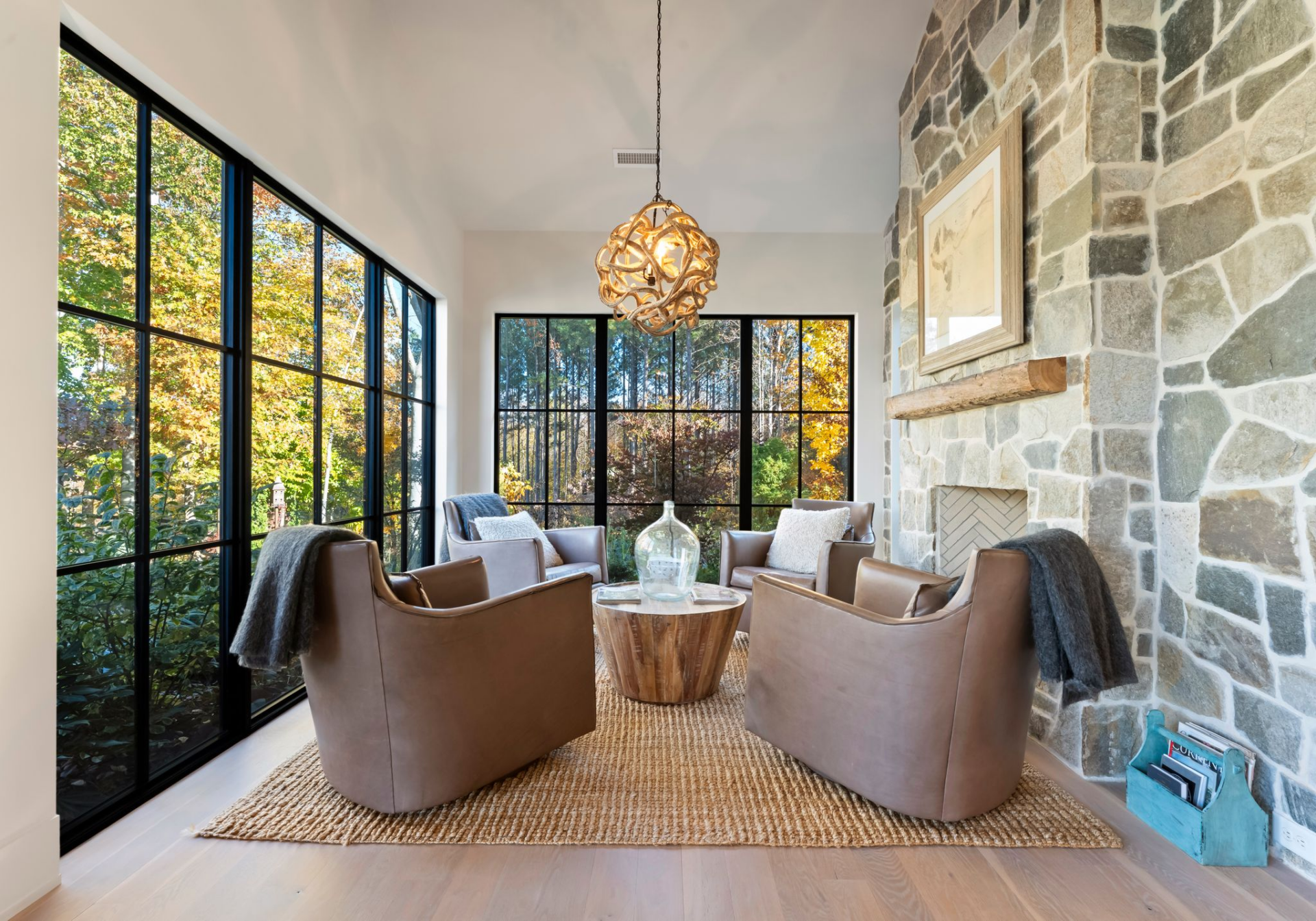 A full stone fireplace wall grounds the space across from floor-to-ceiling black-trimmed windows and adds more texture, while four leather club chairs gather around a round wood table on a woven rug.
