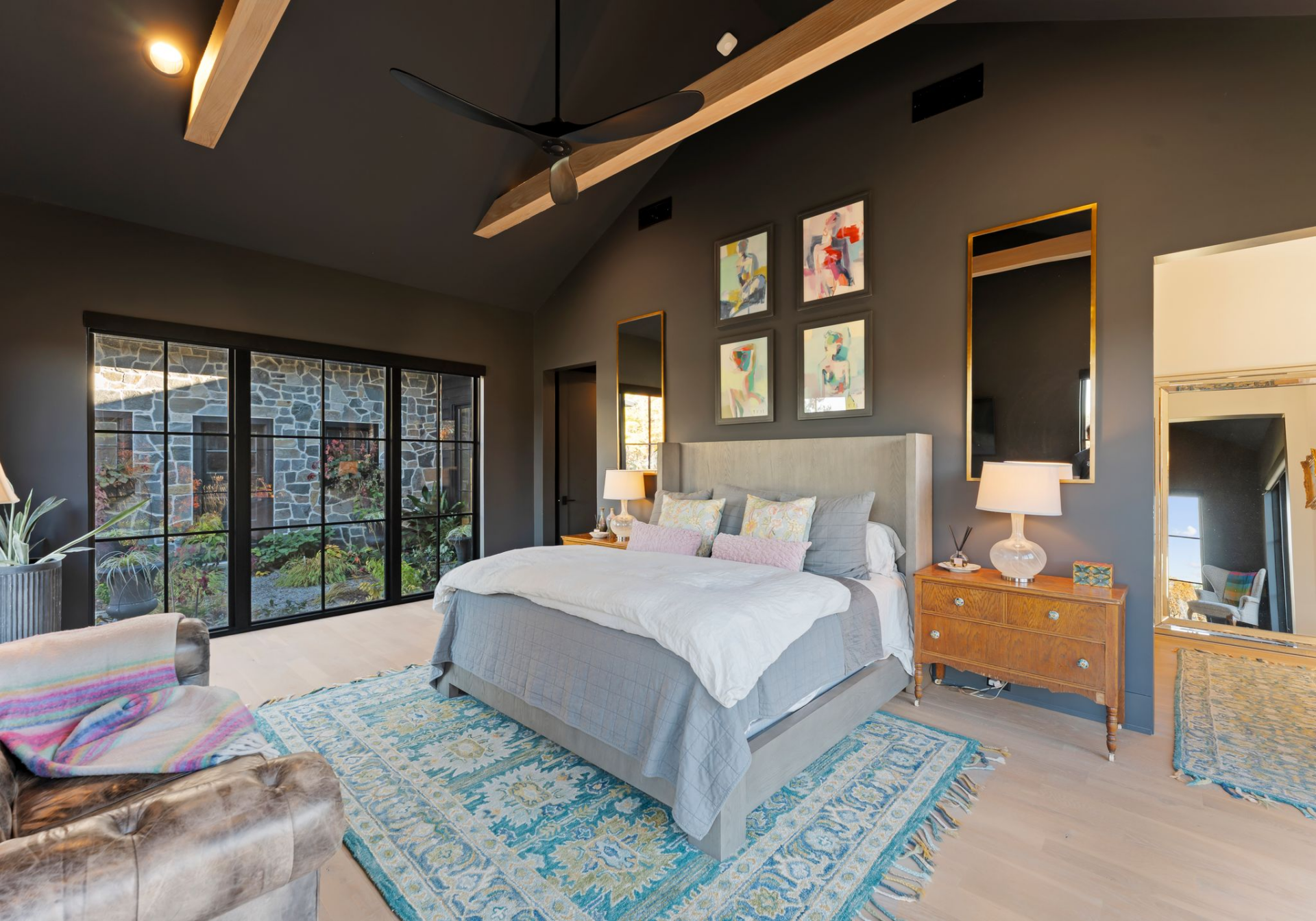 A full wall of black-framed glass looks directly into a garden courtyard, while the layout centers around a simple upholstered bed, framed by wood nightstands and tall mirrors.
