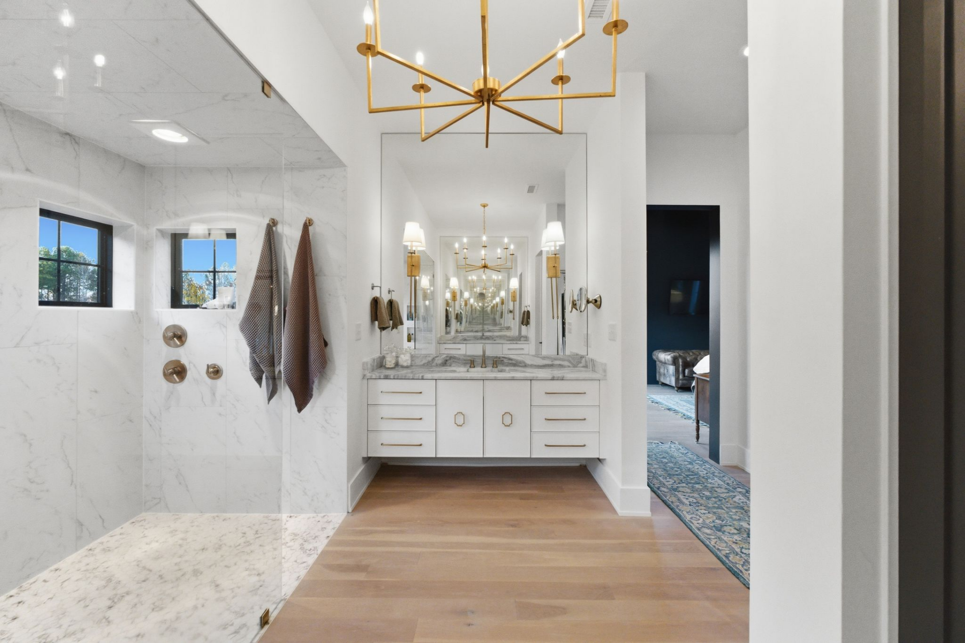 A wide double vanity sits opposite a large walk-in shower, which stretches the width of the room and is enclosed in glass with multiple fixtures and built-in niches. 