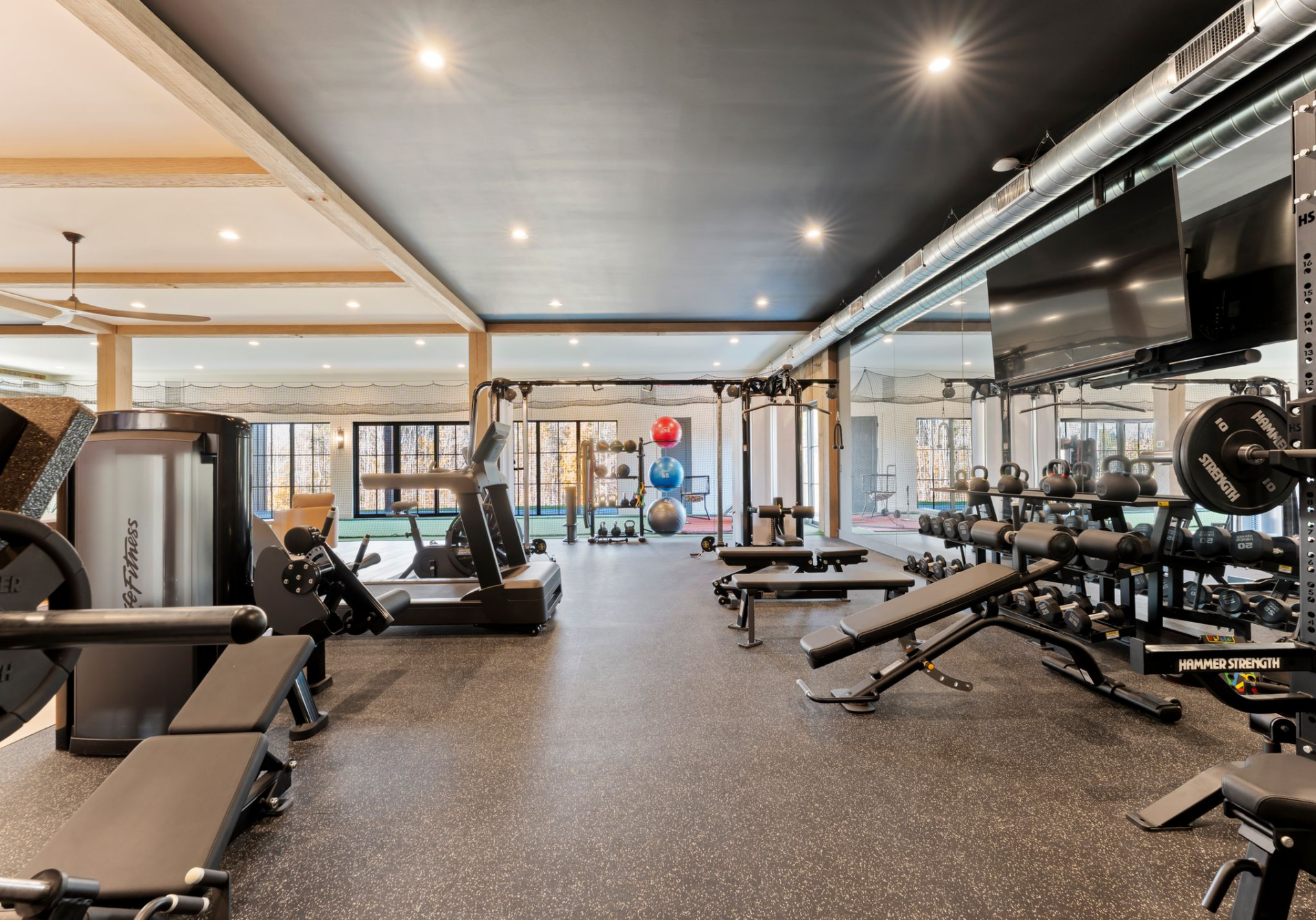 On one side, rubber flooring defines the workout zone, equipped with weight machines, free weights, and cardio equipment, all positioned in front of a mirrored wall. 