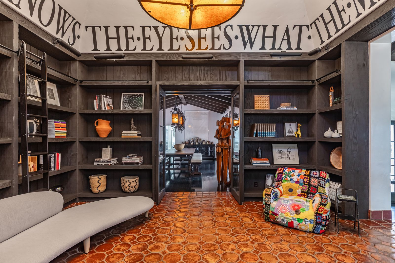 Dark wood built-ins wrap the room from floor to ceiling, filled with books, ceramics, and collected objects atop terracotta tile flooring that guides the eye toward the openings. 